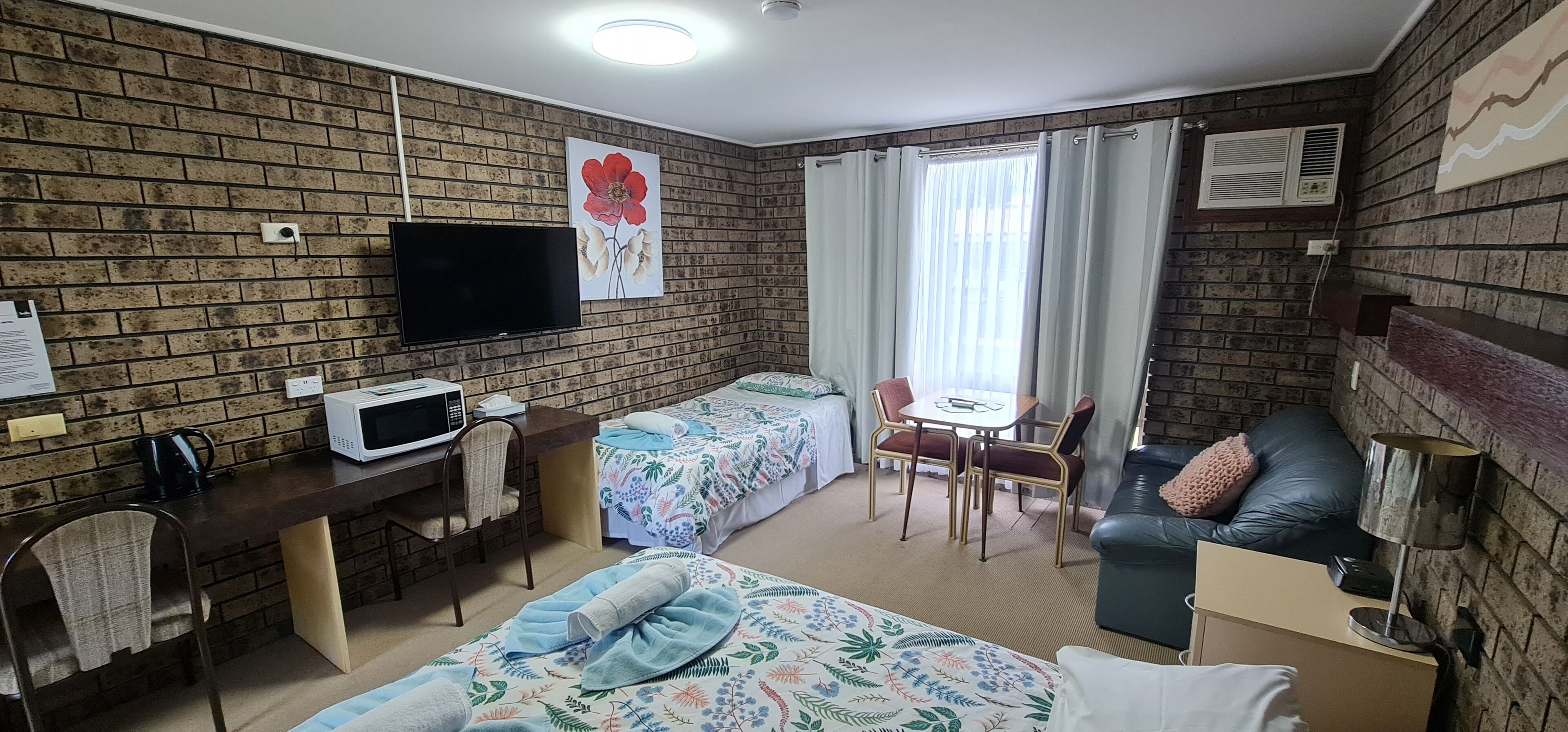 Une chambre d’hôtel avec un mur en briques, un téléviseur à écran plat, un bureau avec un micro-ondes, deux lits avec une literie à motifs floraux, une petite table avec des chaises, un canapé en cuir et une fenêtre avec des rideaux.