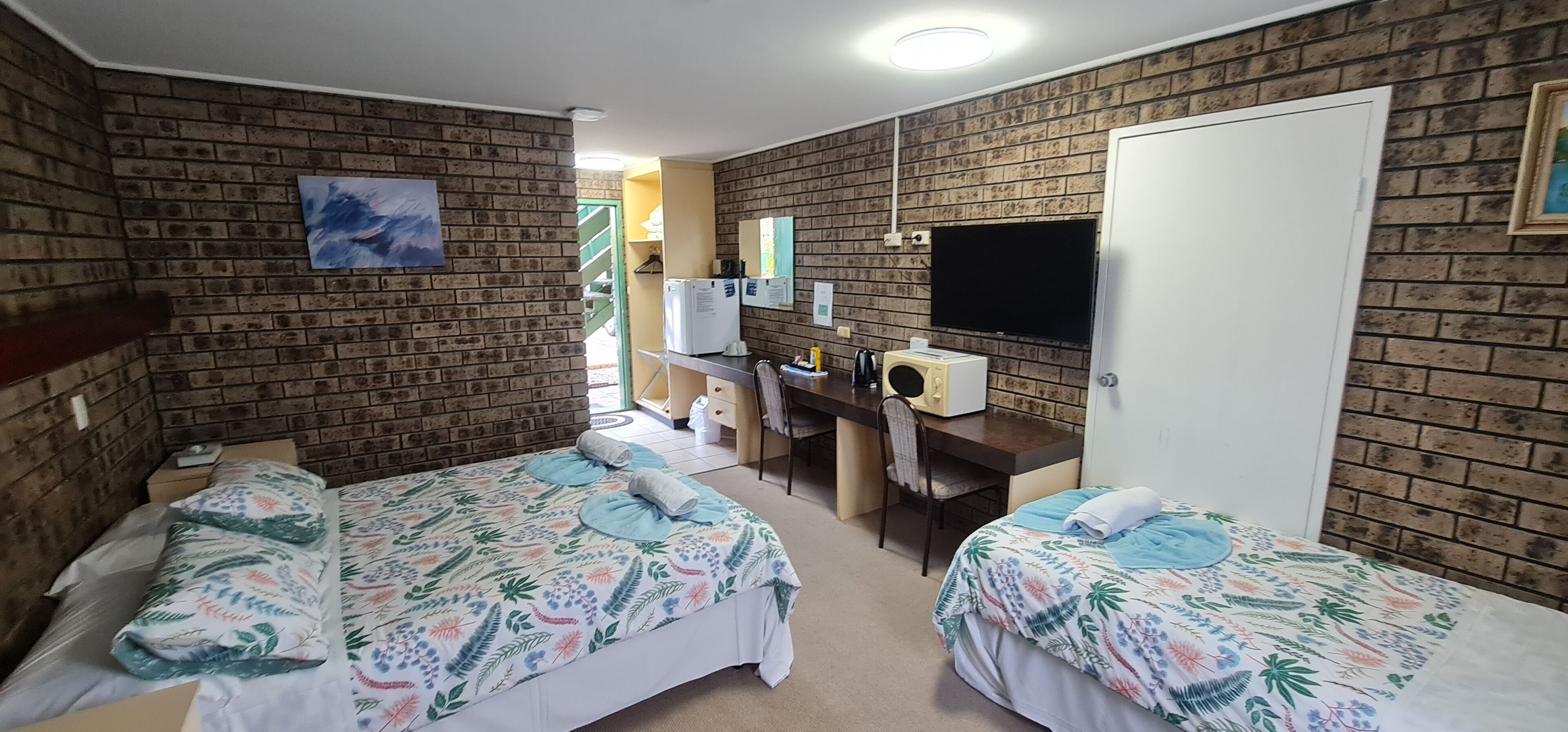 A room with two beds, a desk, a TV, and a brick wall.
