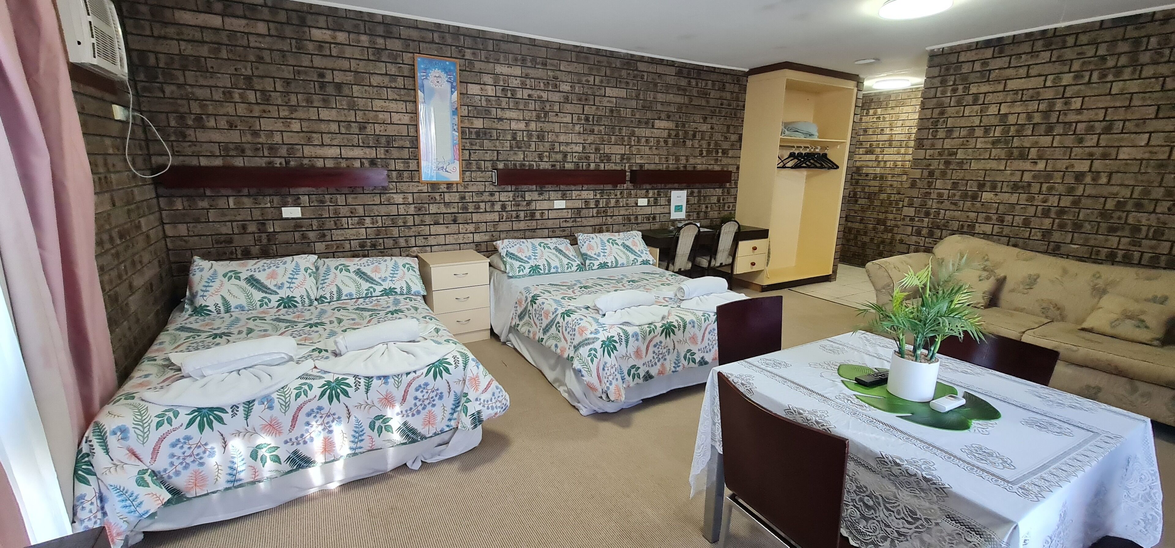 A room with two beds, a sofa, a table with a plant, and a brick wall.
