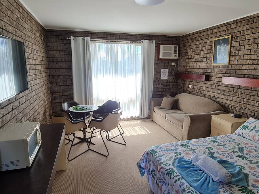 A room with brick walls, a bed with floral bedding, a sofa, a small table with chairs, a window with curtains, and a wall-mounted air conditioner.