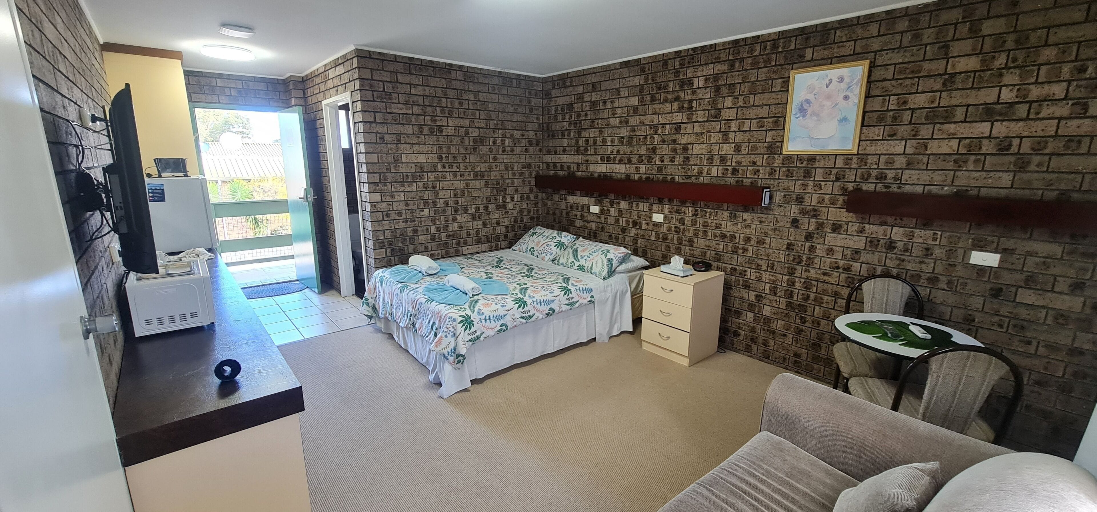A bedroom with a brick-patterned wall, a bed with a floral bedspread, a bedside table, a chair, and a small round table with a green leaf design.