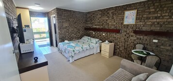 A bedroom with a brick-patterned wall, a bed with a floral bedspread, a bedside table, a chair, and a small round table with a green leaf design.