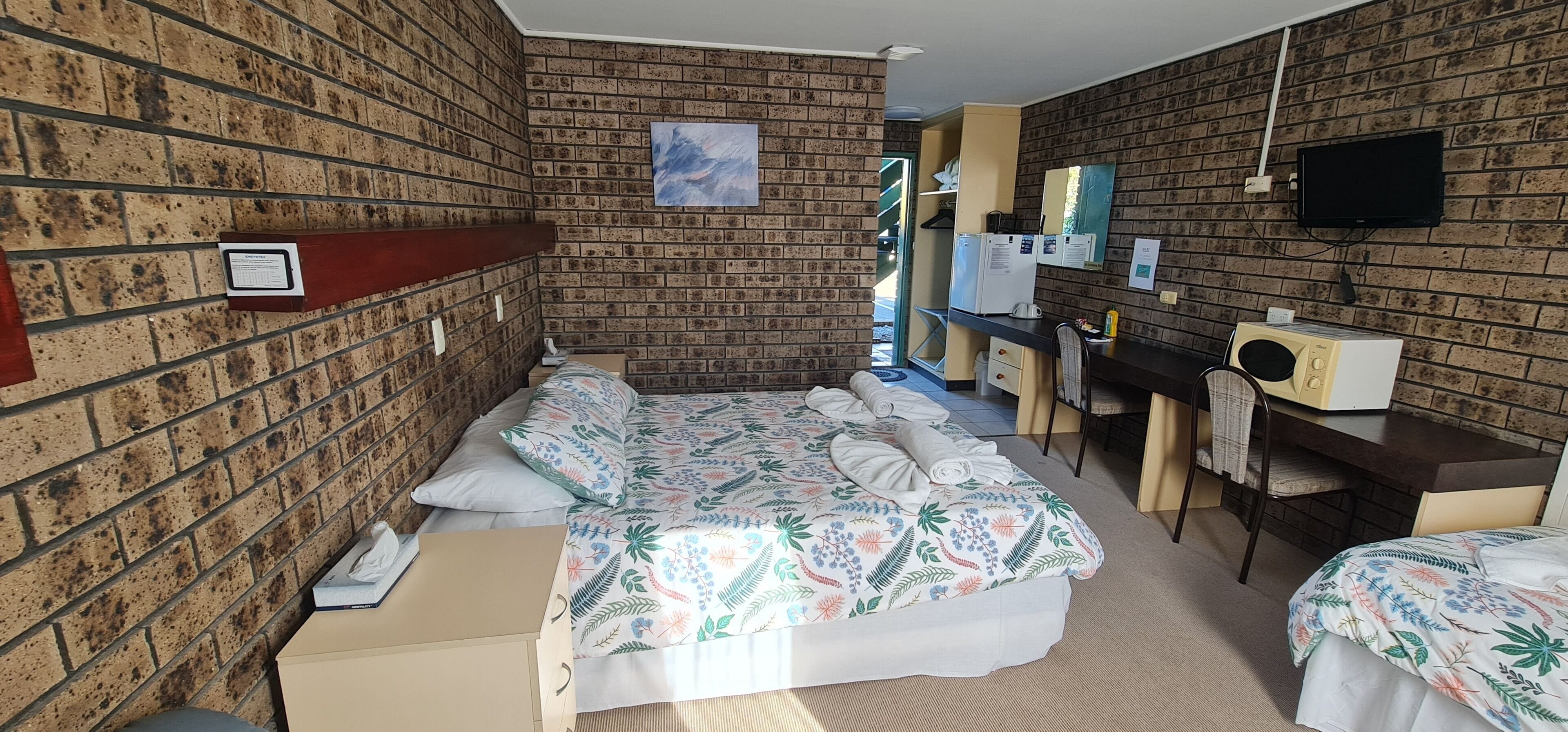 Quarto de hotel com parede de tijolos, cama com roupa de cama florida, escrivaninha com cadeira, televisão e micro-ondas.