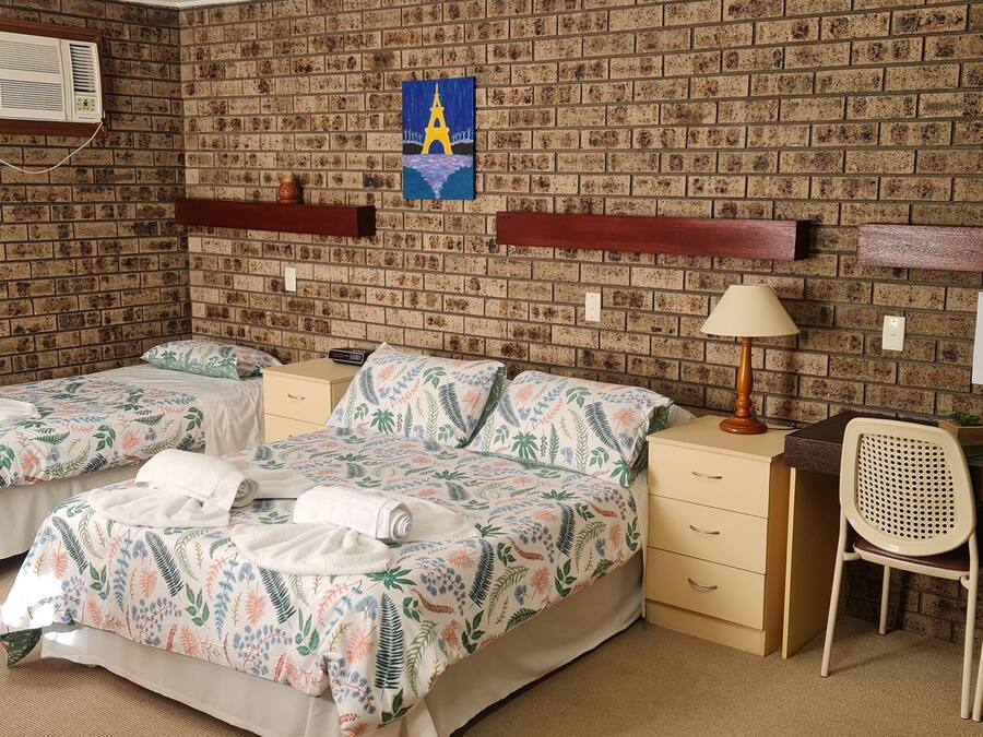 A hotel room with two beds, a desk, two chairs, a lamp, and a brick-patterned wall.