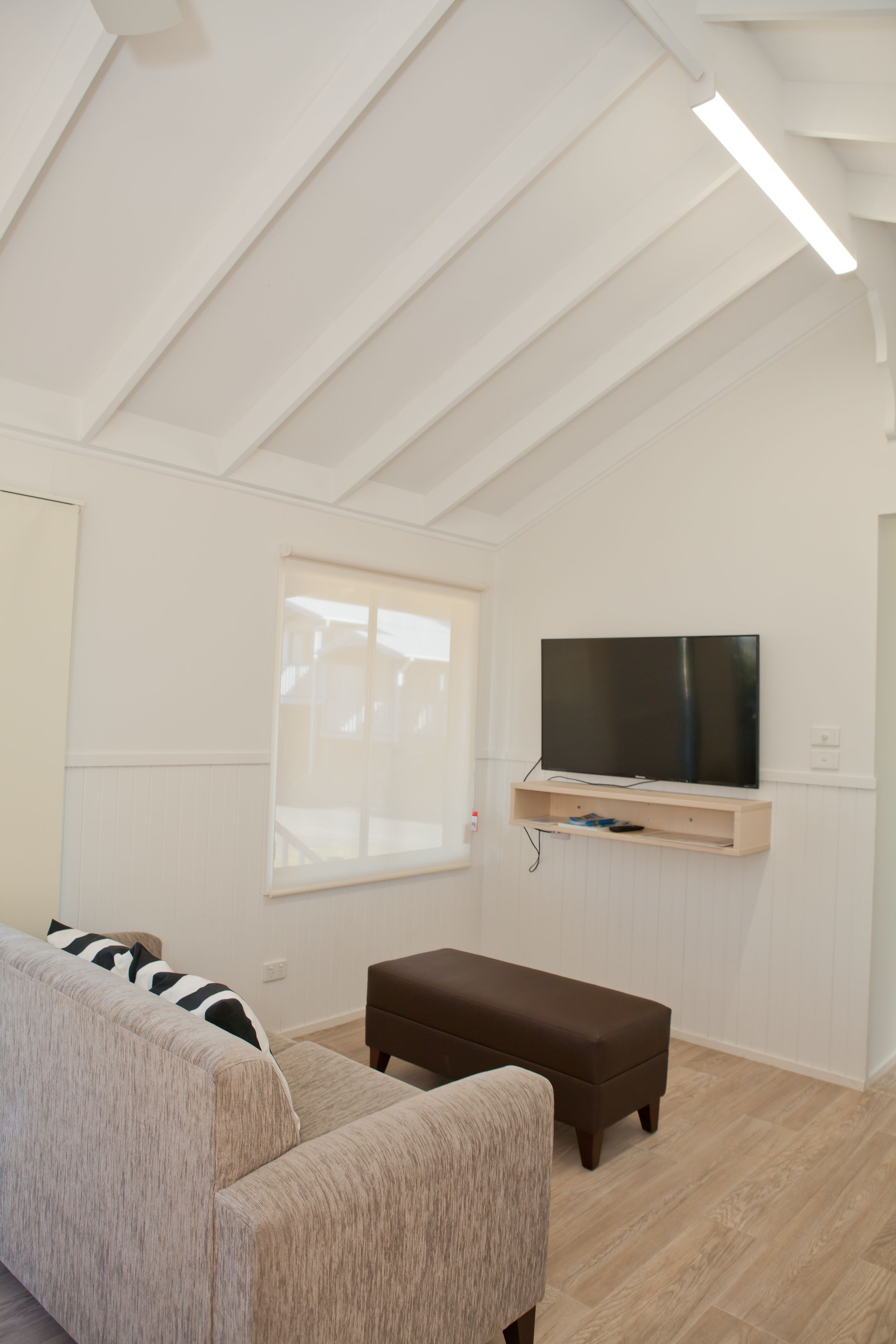 Een moderne woonkamer met een flatscreen televisie aan de muur, een beige bank en een bruin poef.