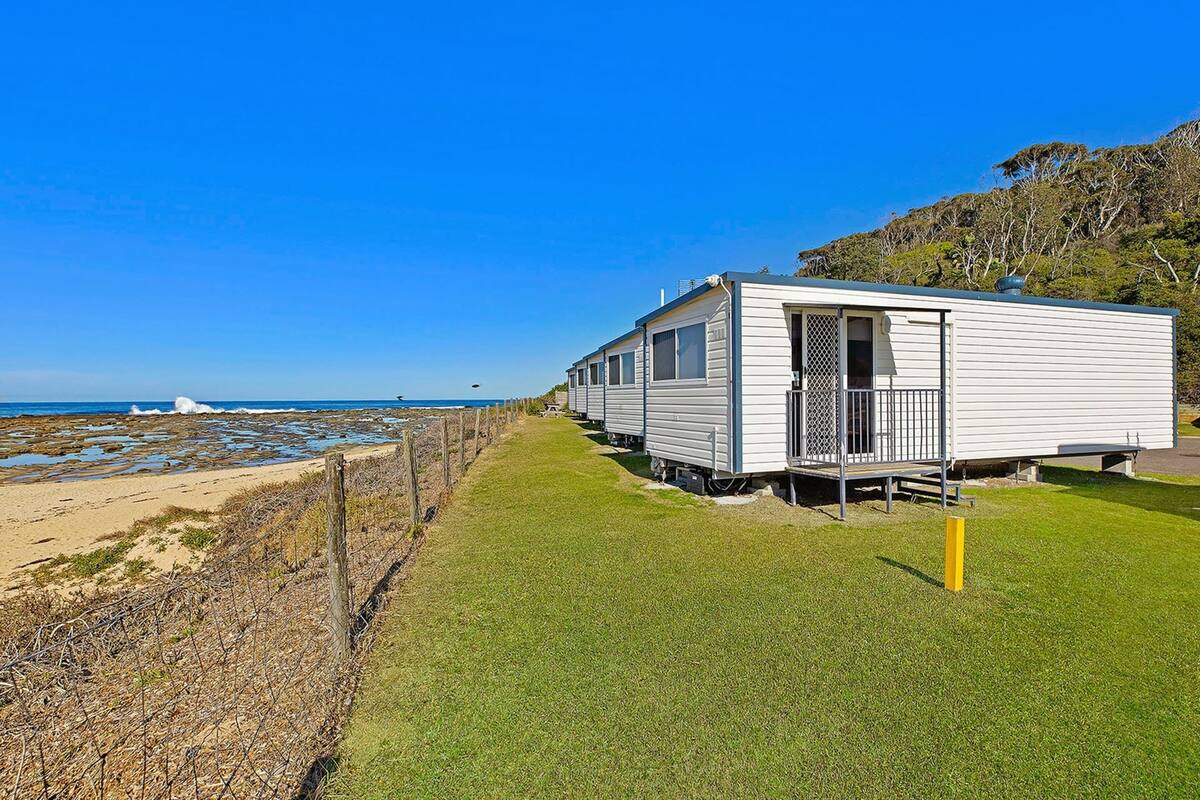 Une rangée de maisons mobiles blanches, avec une pelouse devant et une plage en arrière-plan.