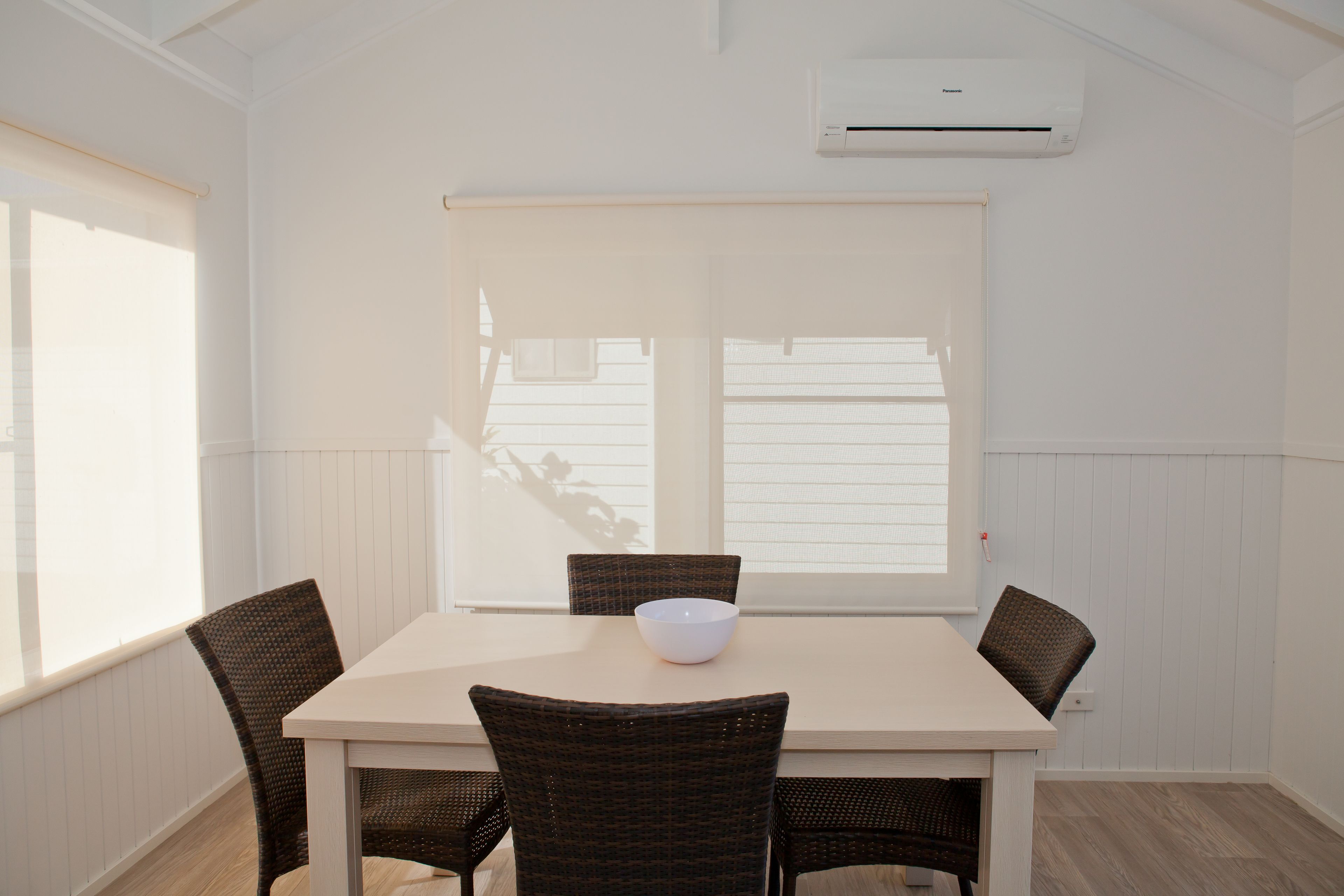 A dining area with a table, four chairs, a bowl, and a window with blinds.
