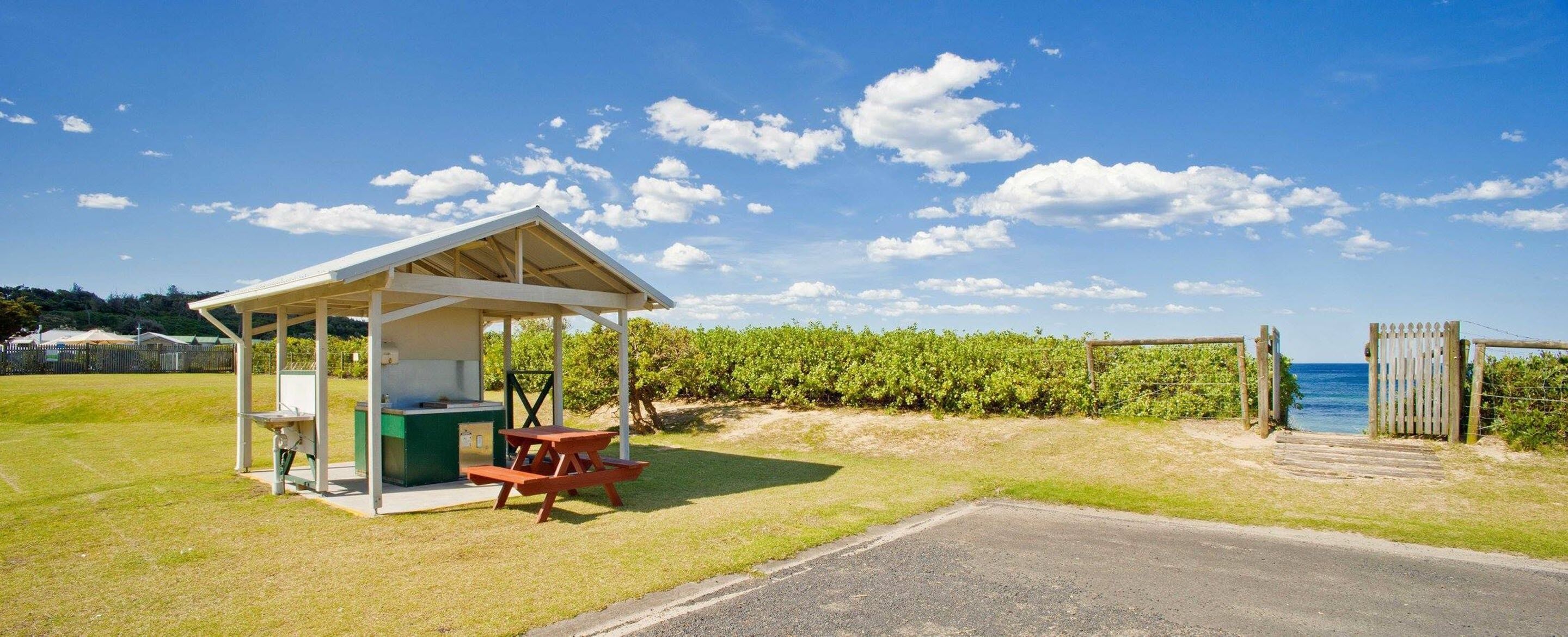 Une petite structure extérieure dotée d’une table de pique-nique et d’un barbecue, située près d’une porte donnant accès à une plage.