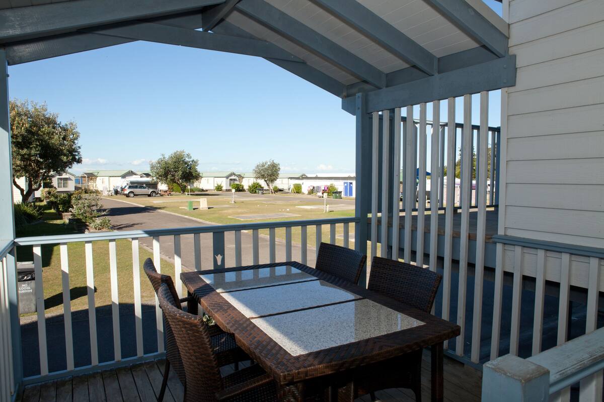 Un espace extérieur abrité, équipé d’une table et de chaises, avec vue sur un parking et un bâtiment en arrière-plan.