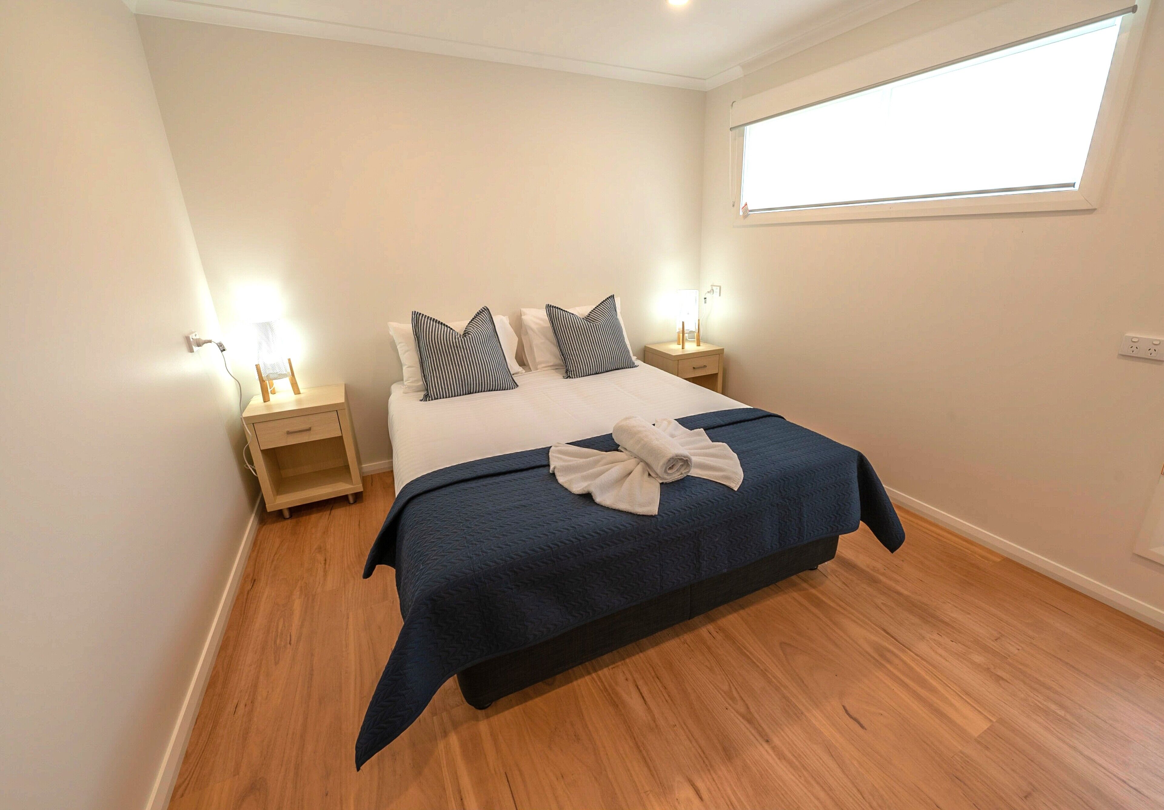 A neatly made bed with a dark blue bedspread and two pillows, a small bedside table with a lamp, and a window providing natural light.