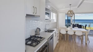 A modern kitchen with white cabinets, a gas stove, and a dining area with a view of the ocean. - Blue Lagoon Beach Resort (Bateau Bay)