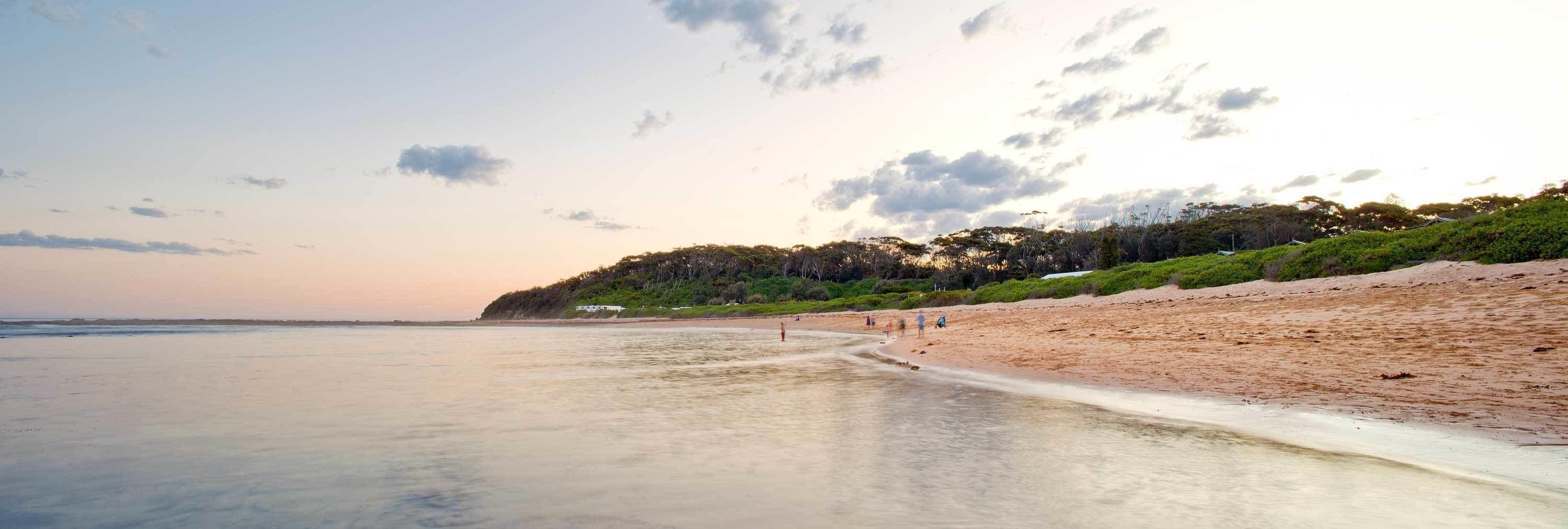 A beach with a clear sky, calm sea, and a sandy shore with greenery on the hillside.