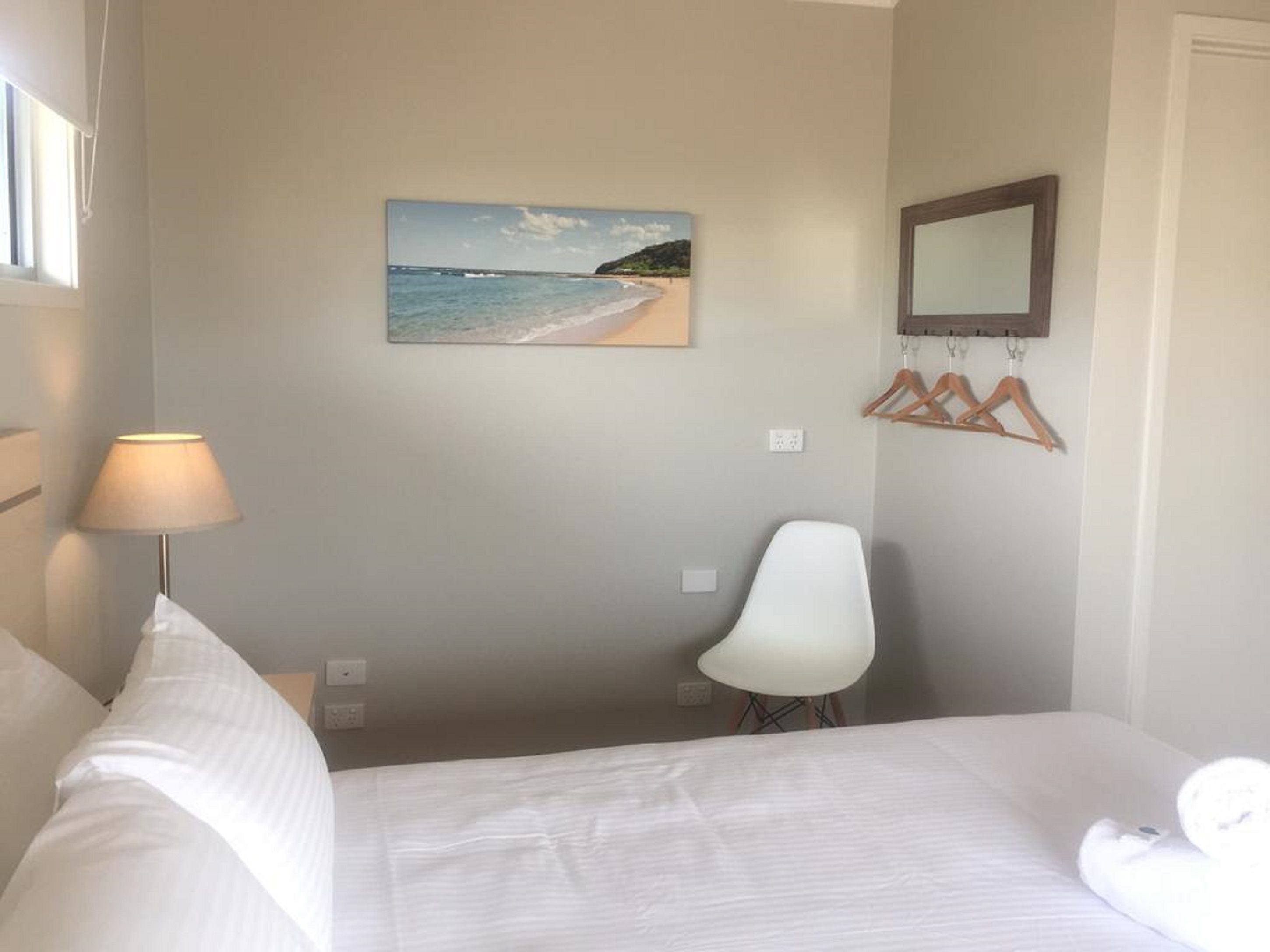 Una habitación de hotel con cama, escritorio, silla, lámpara, un cuadro de playa y espejo en la pared.