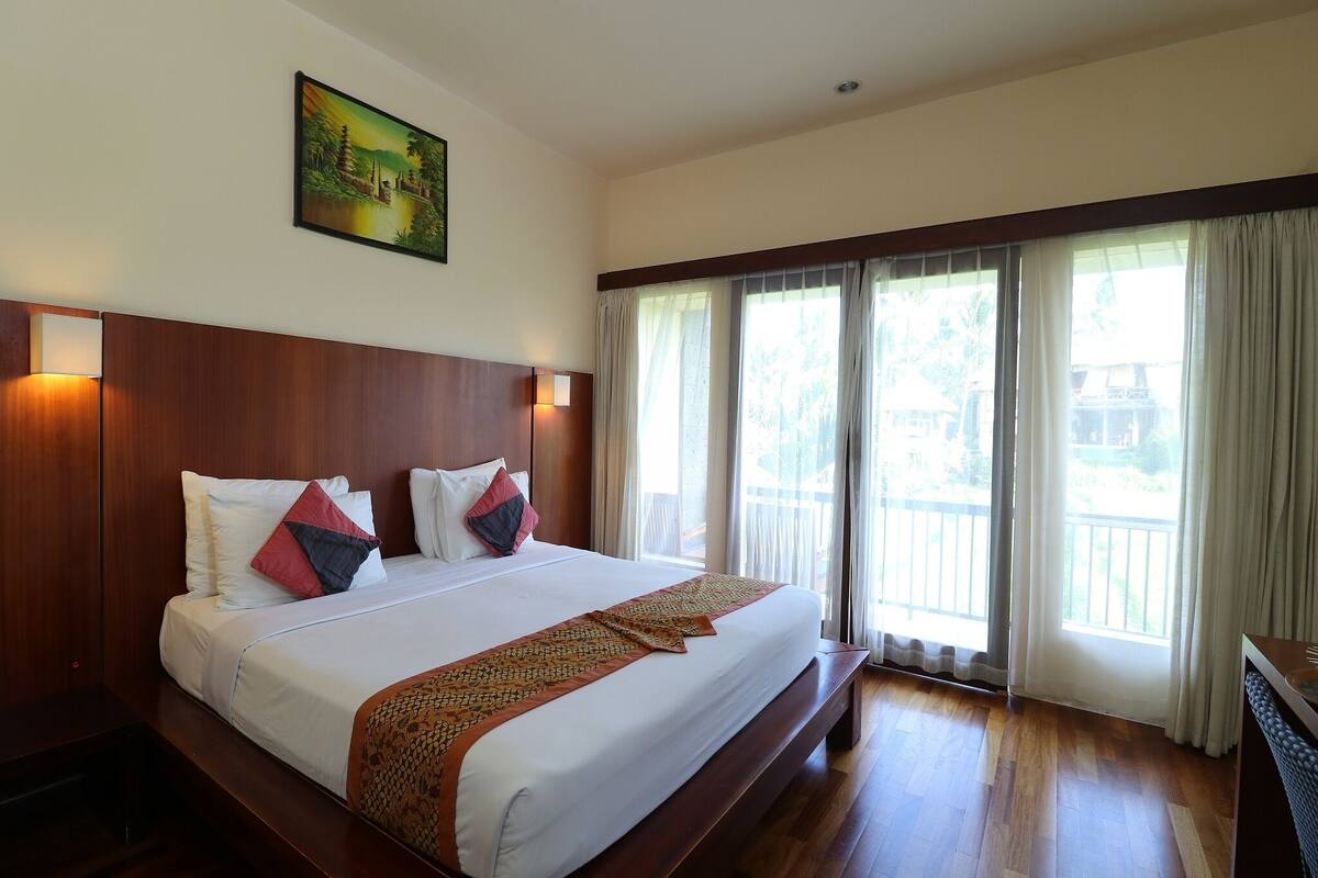 A large bed with a wooden headboard, white bedding, and decorative pillows. There is a painting on the wall above the bed and a window with curtains to the right.