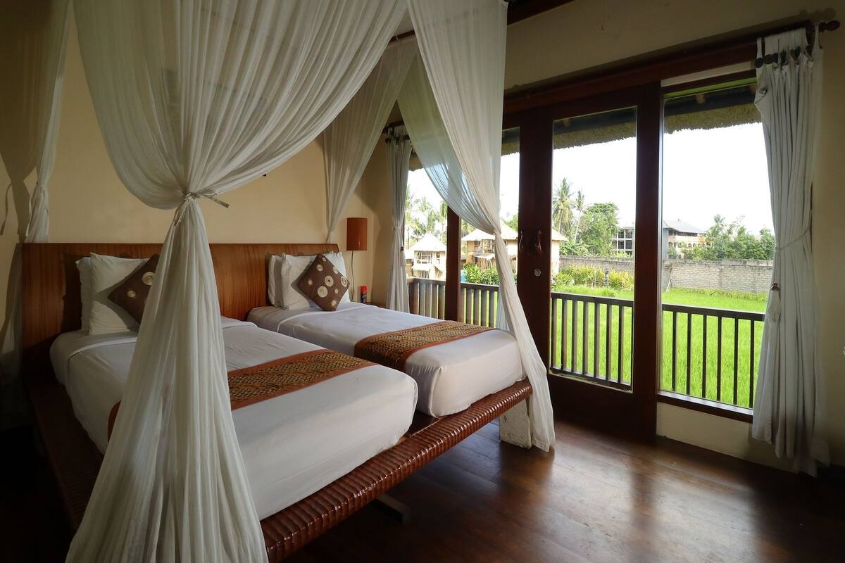 A bedroom with two beds, a wooden headboard, and a balcony with a view of the greenery and buildings.