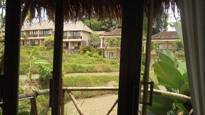 Vista desde un espacio al aire libre techado con barandas de bambú, una silla y una vista de un complejo turístico con varias construcciones y abundante vegetación.