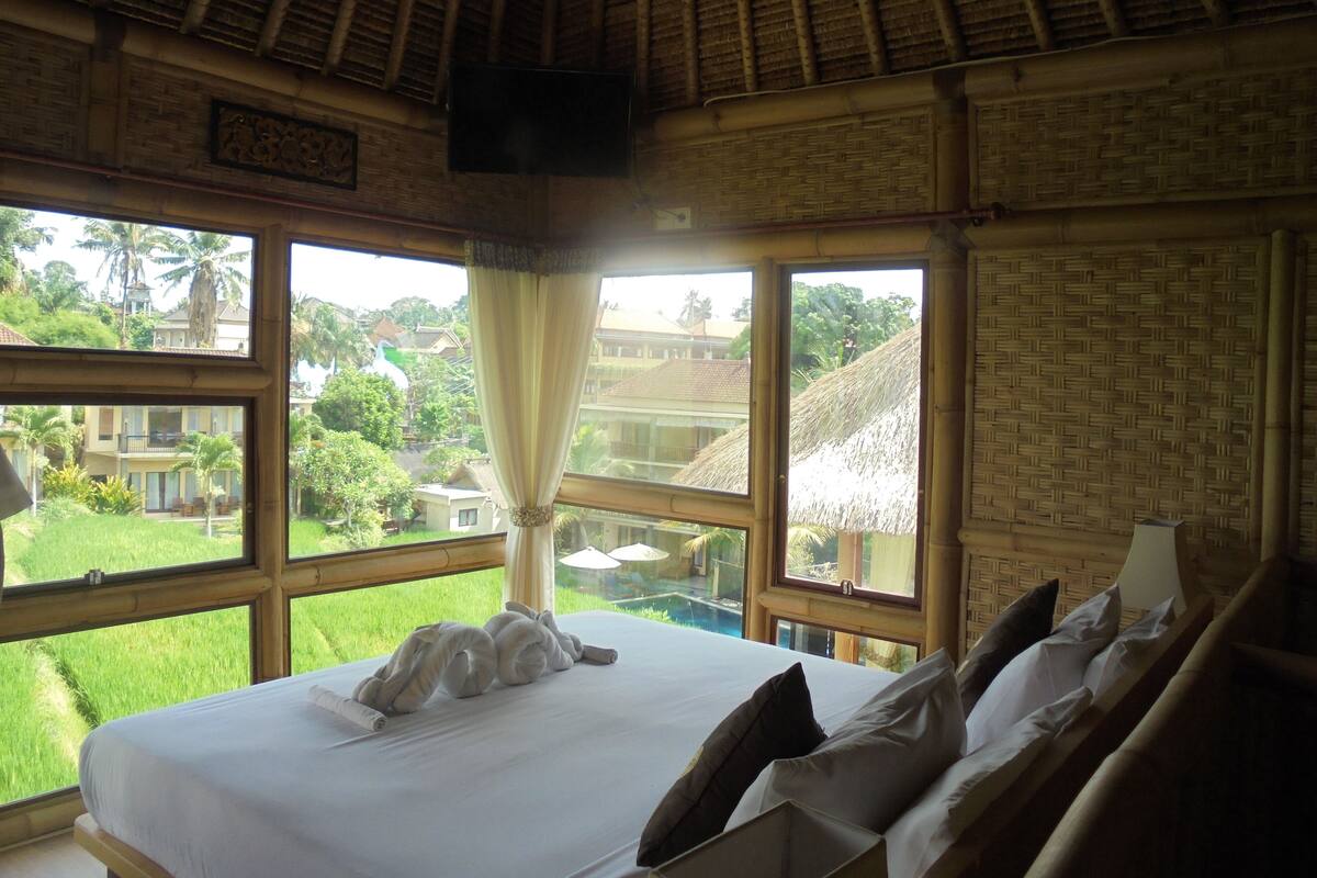 A hotel room with a bed, pillows, a curtain, and a view of a pool and greenery outside.
