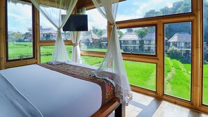 Un dormitorio con cama, televisor y ventanas amplias que ofrecen vistas a campos verdes y casas.