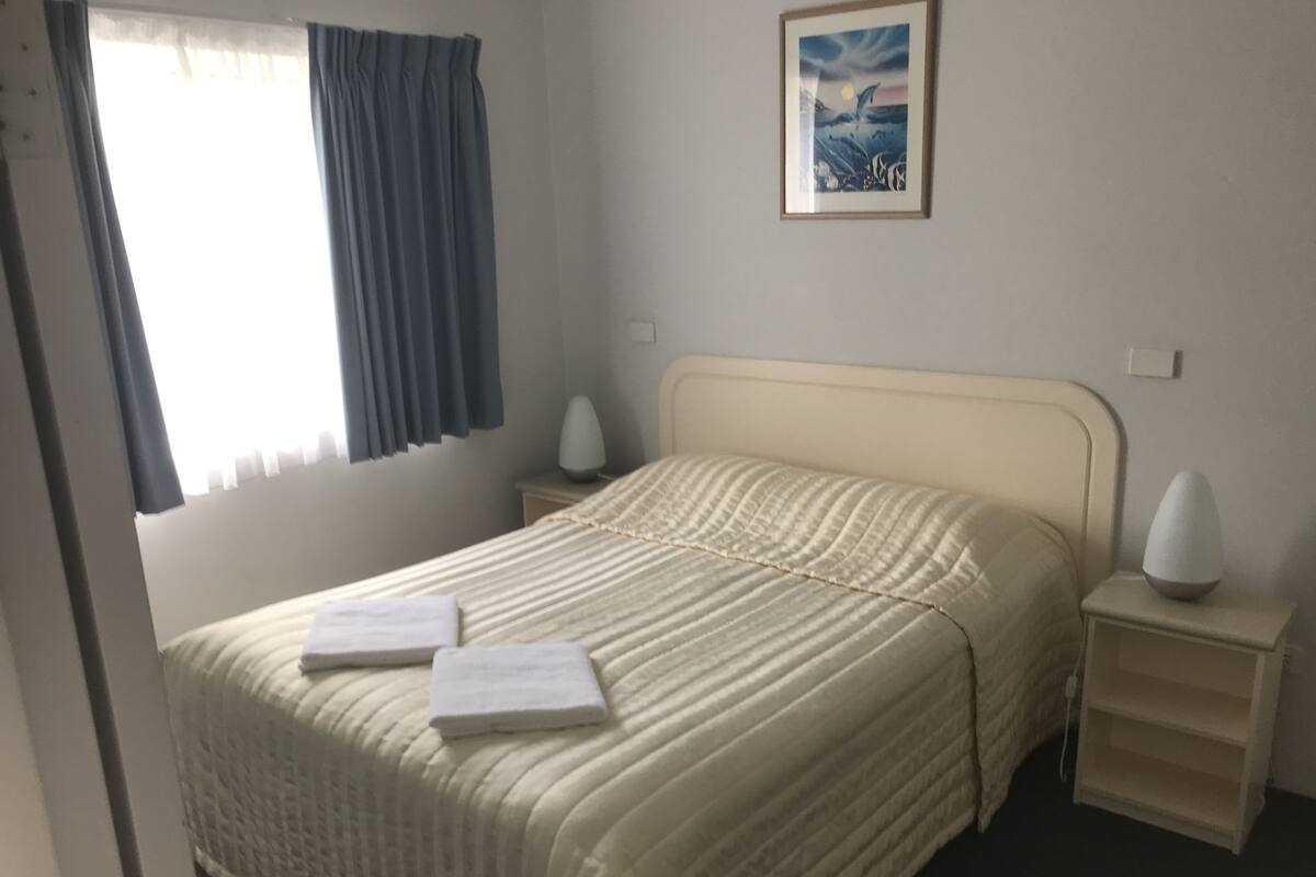 A neatly made bed with a striped bedspread and two pillows, a bedside table with a lamp, a window with curtains, and a framed picture on the wall.