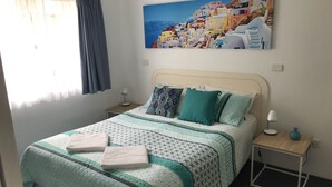 Una cama bien hecha con una colcha estampada, dos almohadas y dos toallas dobladas. Hay una mesita de noche con una lámpara y un florero. Sobre la cama cuelga un cuadro de un pueblo costero.