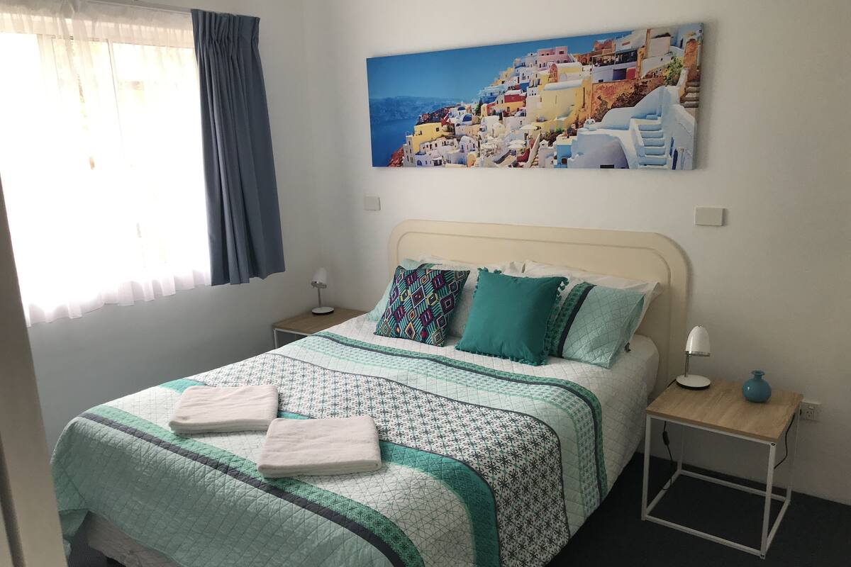 A neatly made bed with a patterned bedspread, two pillows, and two folded towels. There is a bedside table with a lamp and a vase. A painting of a coastal village hangs above the bed.