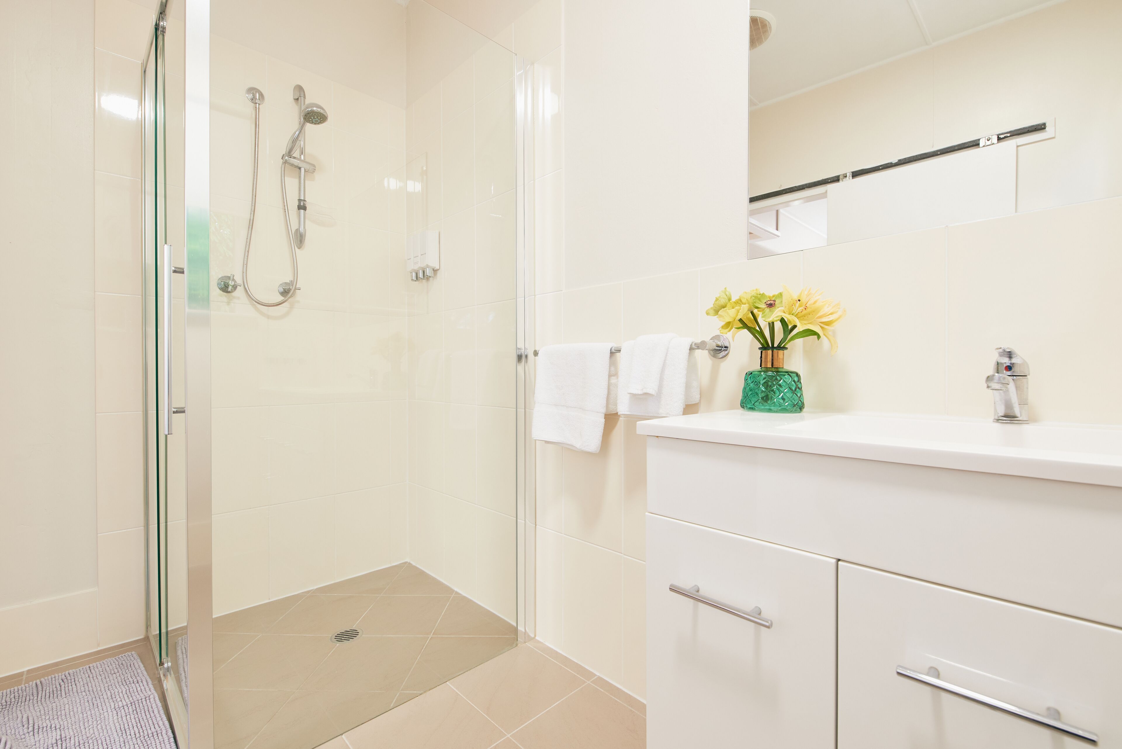Un baño moderno con cabina de ducha de vidrio, tocador blanco y un florero verde, y un perchero para toallas.