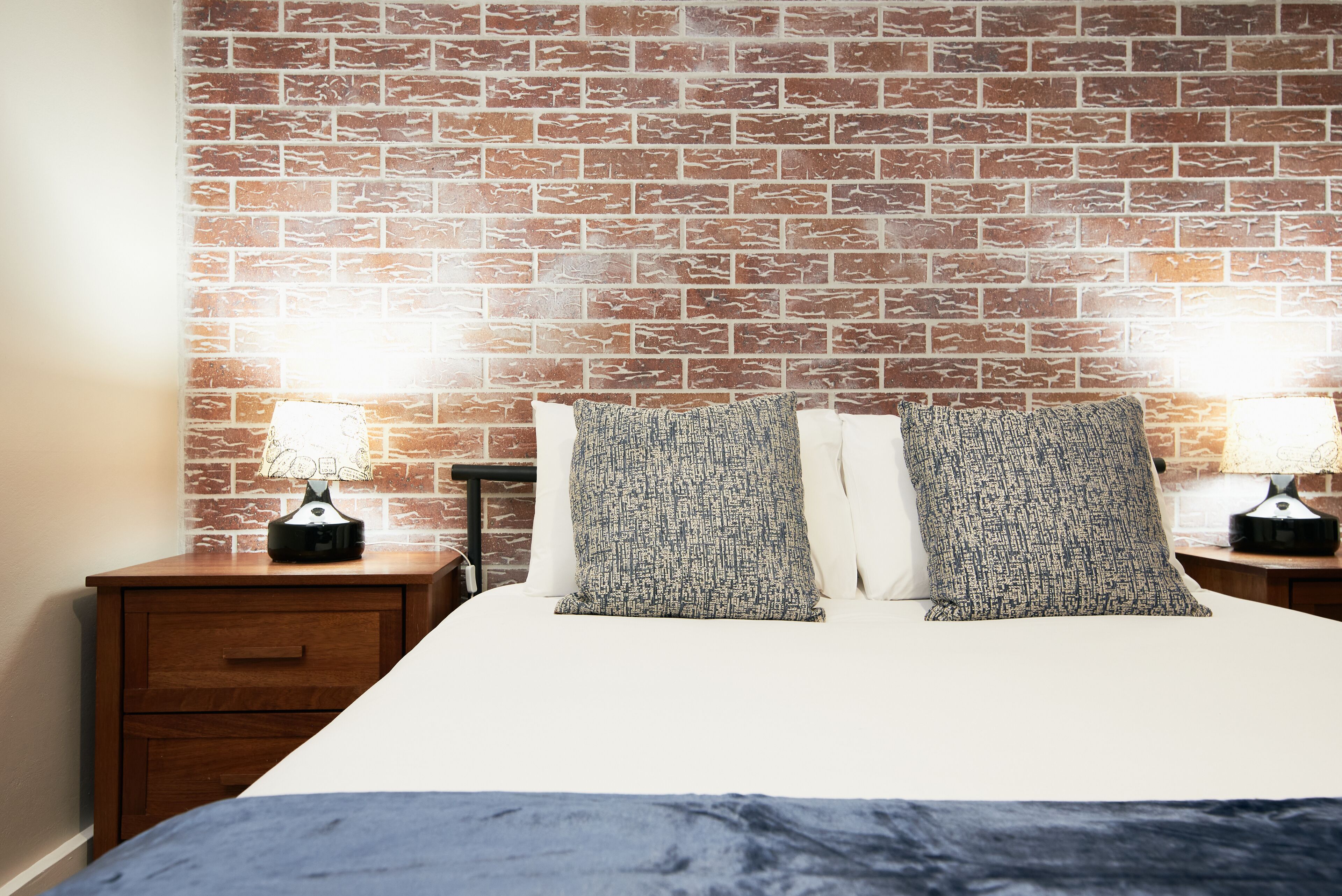 Een slaapkamer met een wand met baksteenmotief, een bed met twee kussens, een nachtkastje met een lamp en een houten lade.
