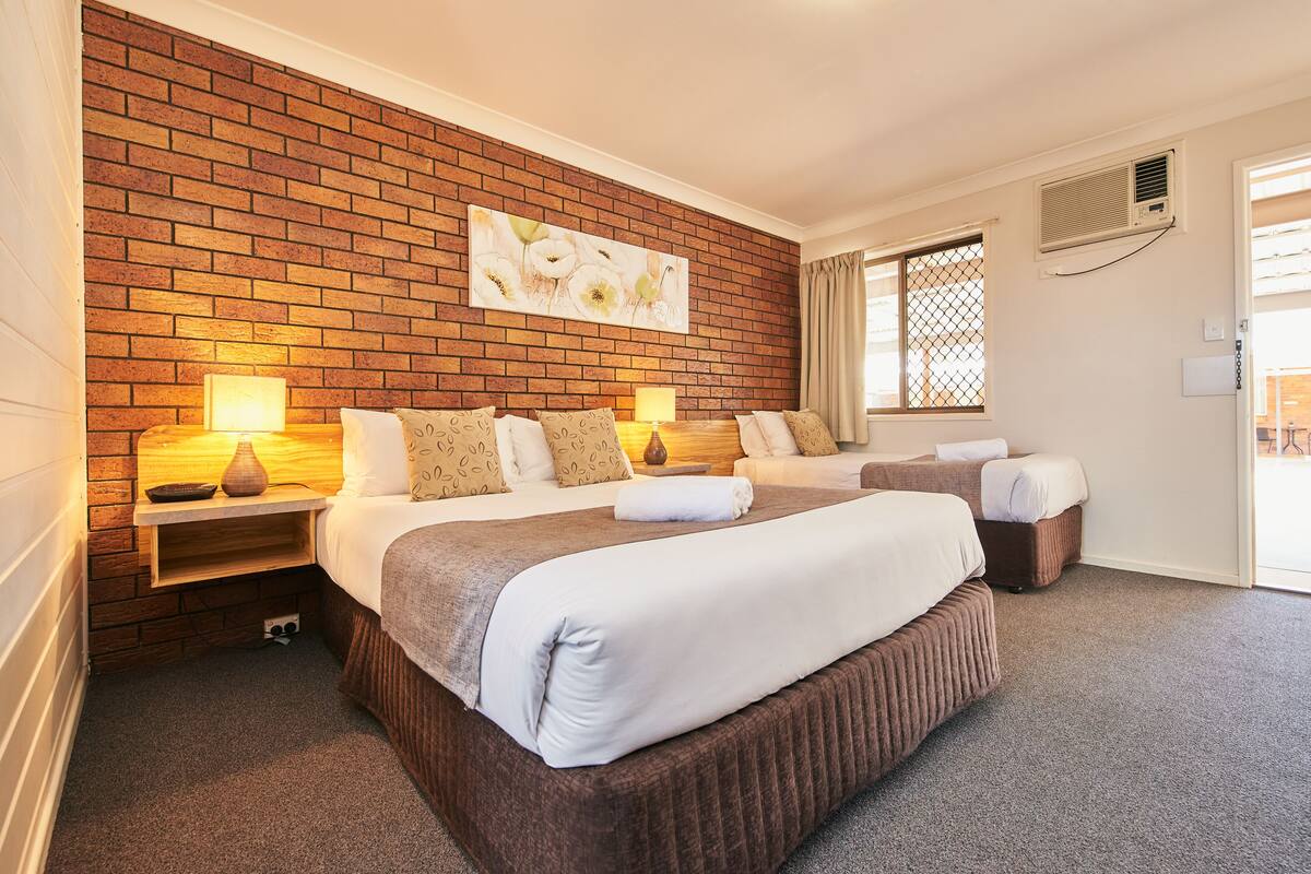 Habitación de hotel con pared de ladrillo, dos camas, lámparas de noche, una obra de arte floral grande y una ventana con cortinas.