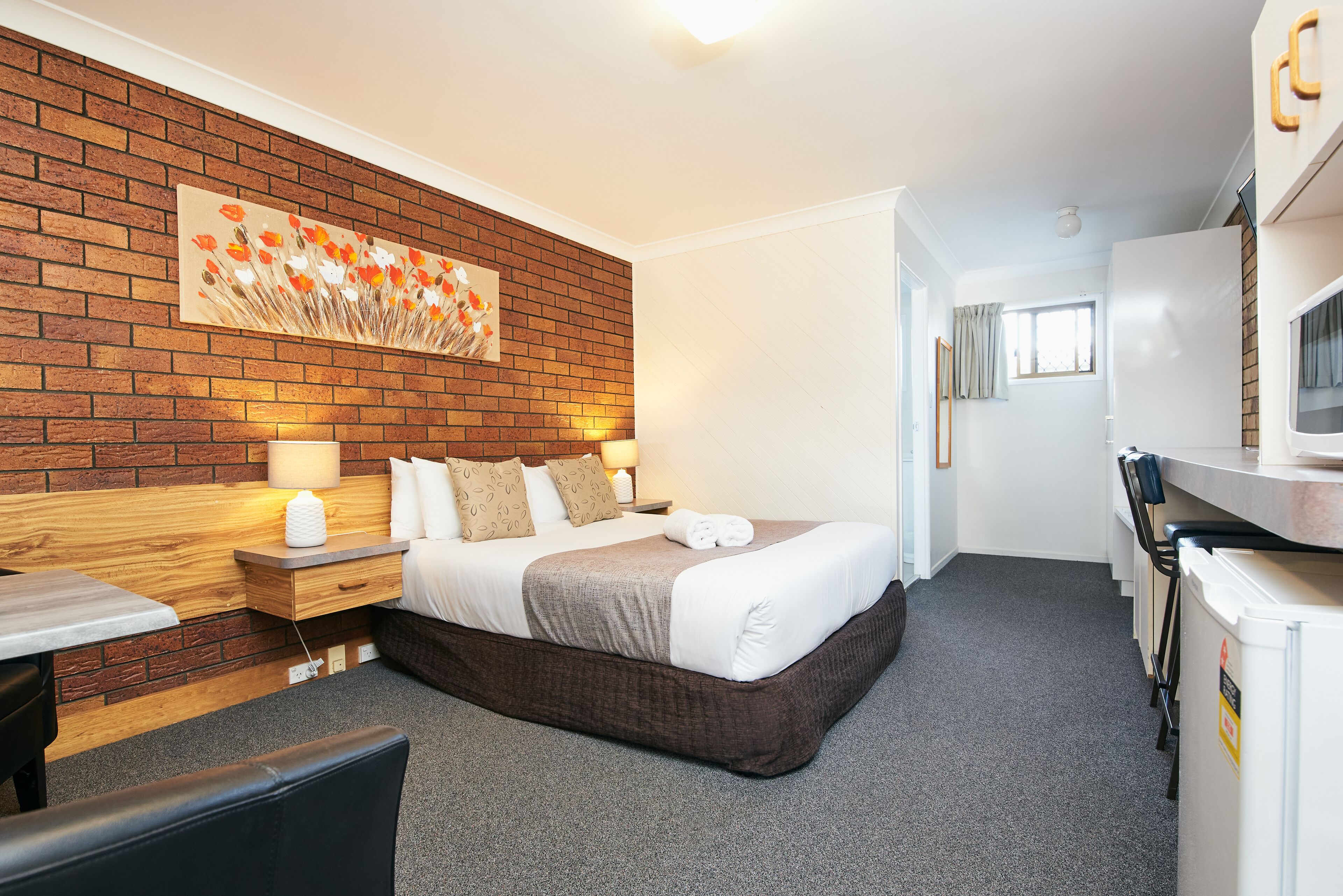 A hotel room with a brick wall, a bed with white linens, a wooden nightstand, a desk, and a kitchenette.