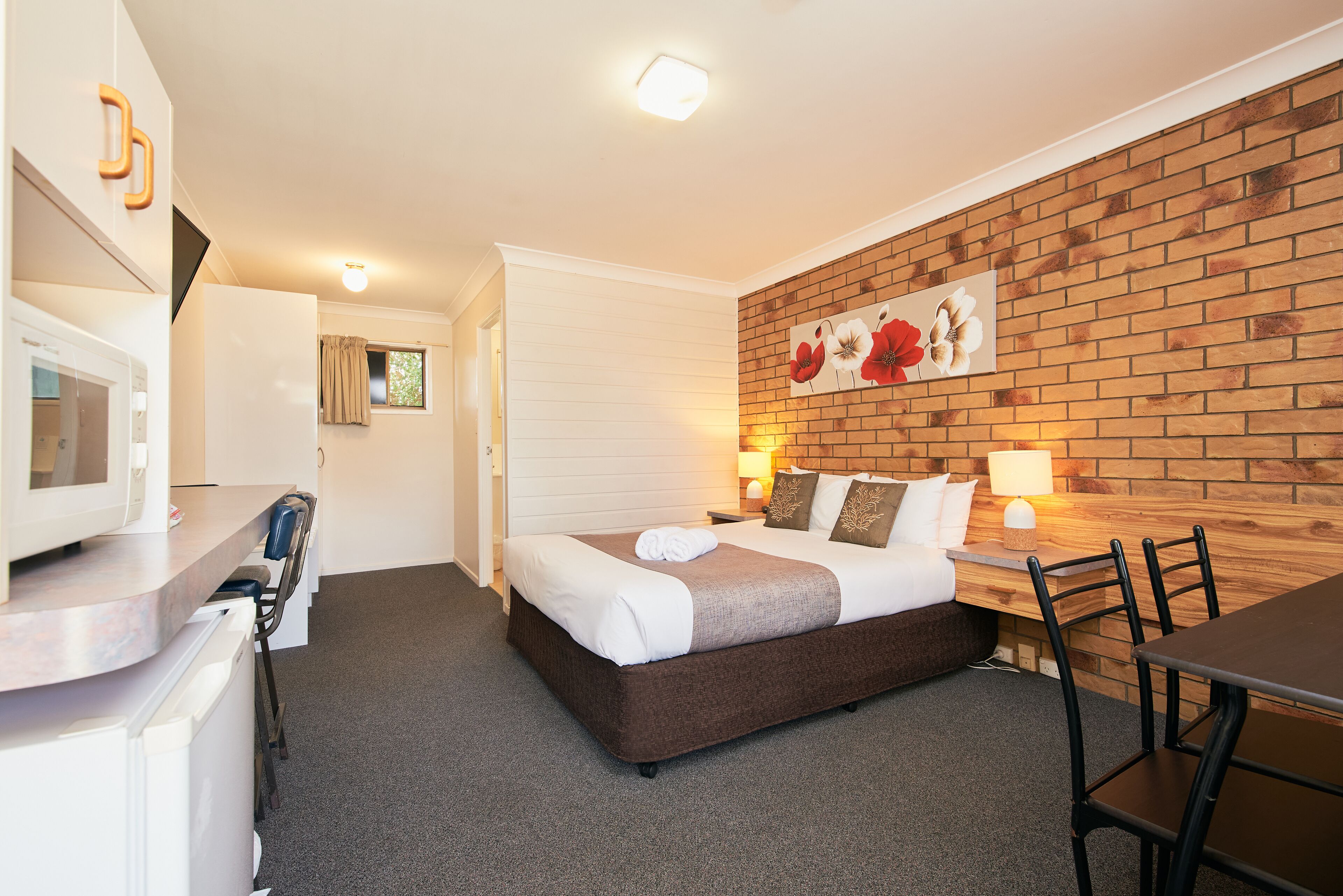 A hotel room with a bed, a desk, a microwave, a chair, and a brick wall with a floral painting.