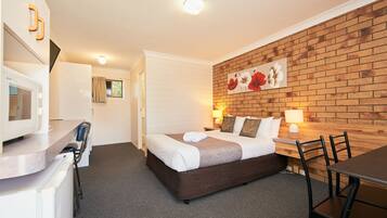A hotel room with a bed, a desk, a microwave, a chair, and a brick wall with a floral painting.