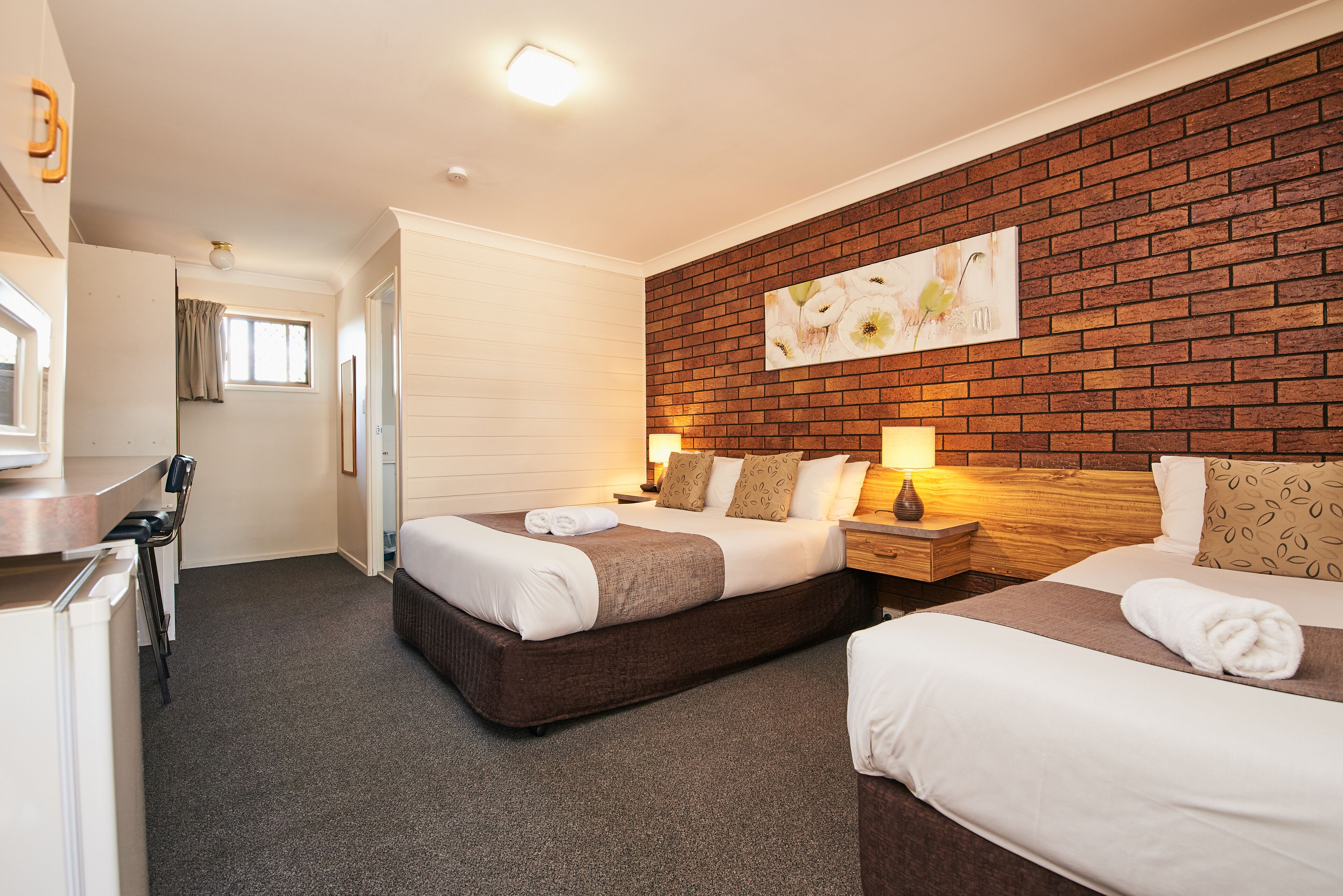 A hotel room with two beds, a brick wall, a painting, and a desk with a chair.