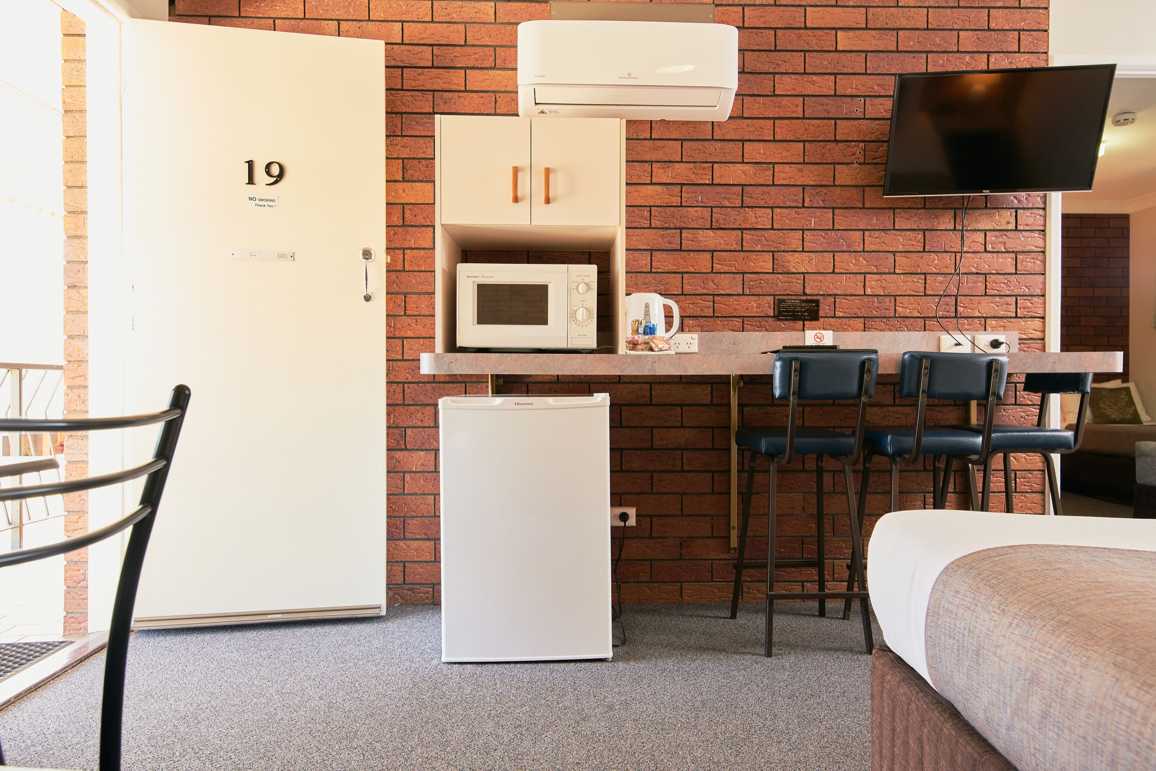 A compact hotel room with a kitchenette, a single bed, a TV mounted on the wall, and a brick-patterned wall.