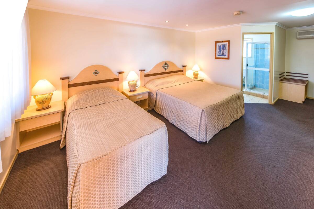 A hotel room with two beds, each with a headboard and bedside tables. There is a door leading to a bathroom with a shower.