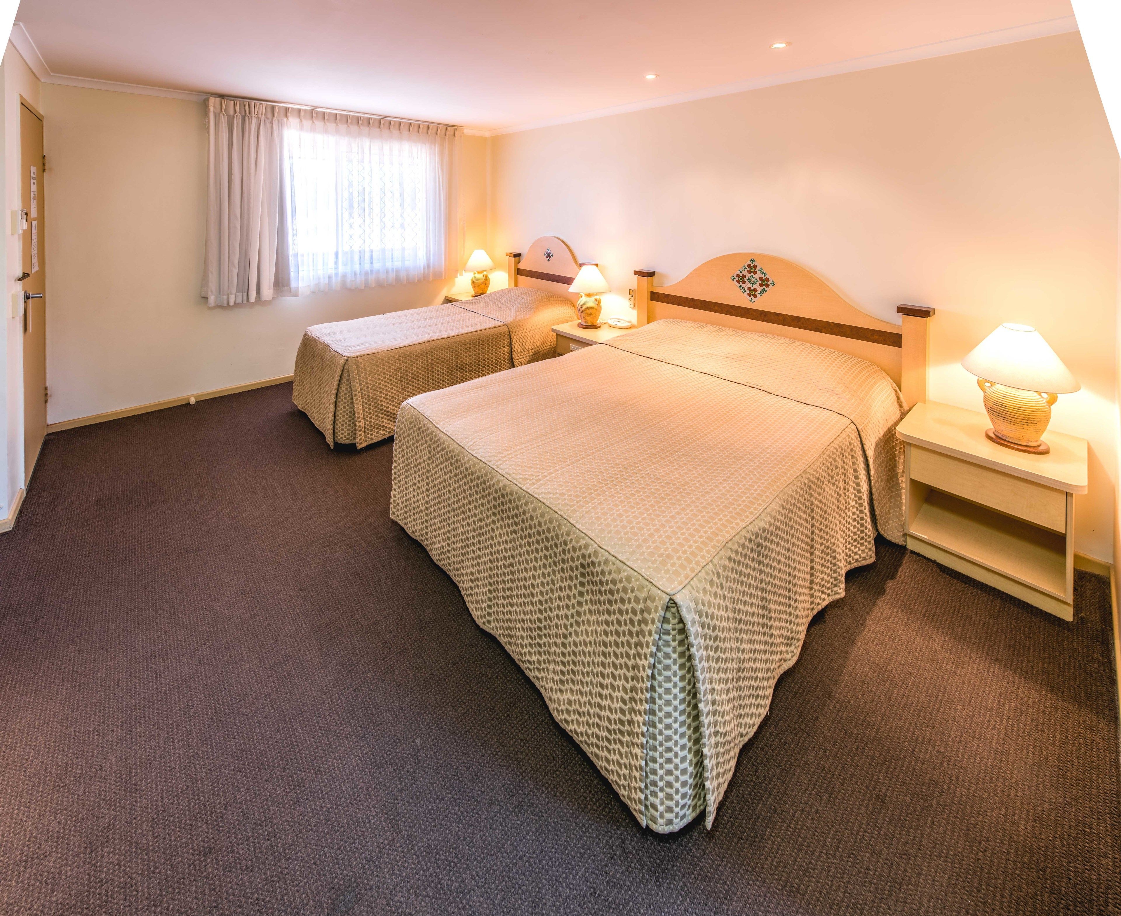 A hotel room with two beds, a nightstand with a lamp, a window with curtains, and a door with a lock.