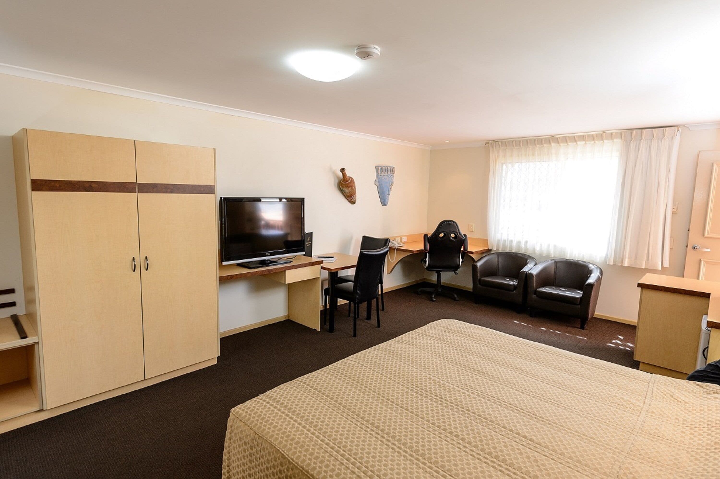 Quarto de hotel com uma cama, uma mesa com computador, televisão, guarda-roupa e duas cadeiras.