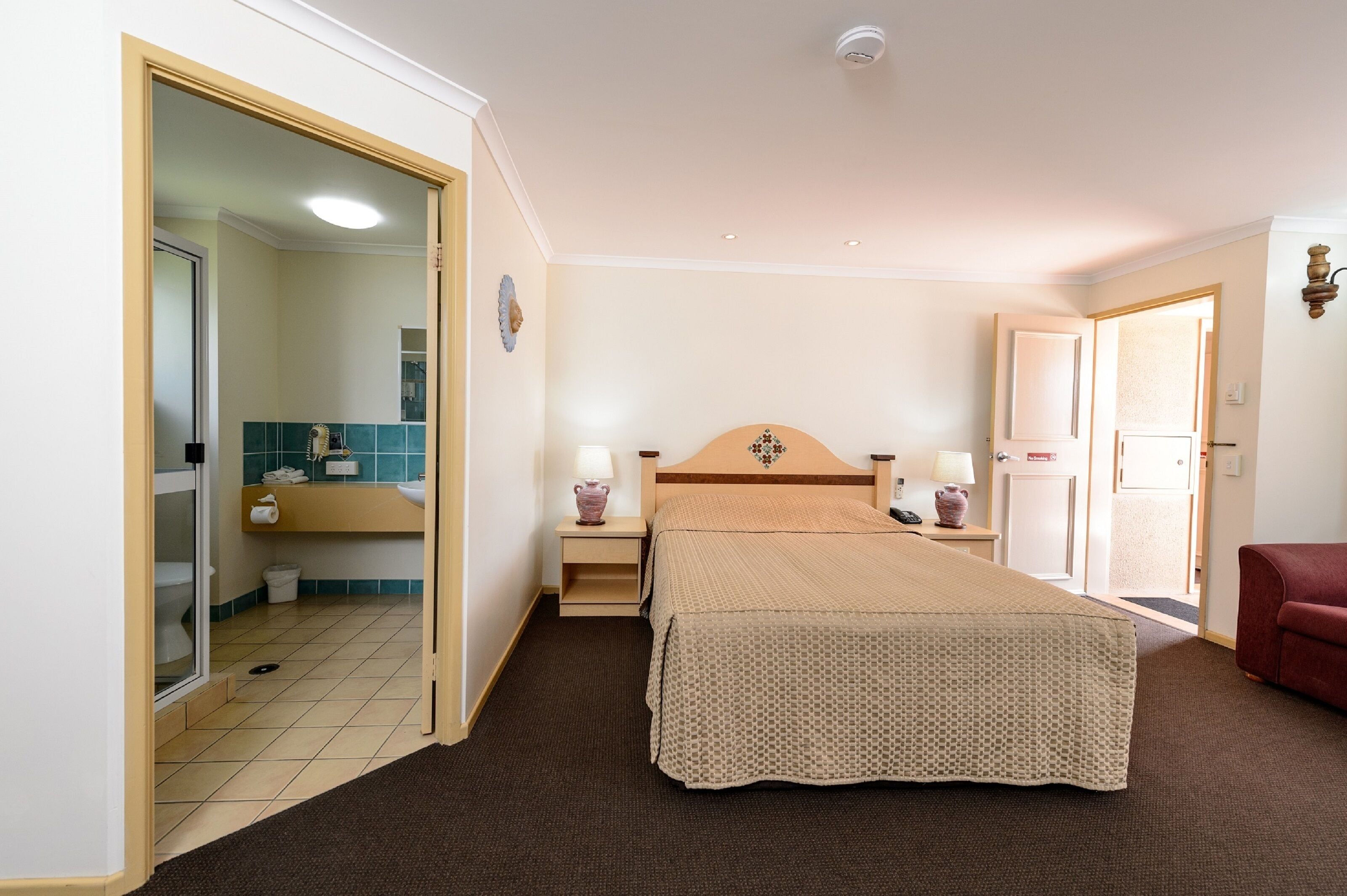 Une chambre d’hôtel avec un lit, des tables de chevet, un miroir, une salle de bain avec un lavabo et des toilettes, et une chaise rouge.