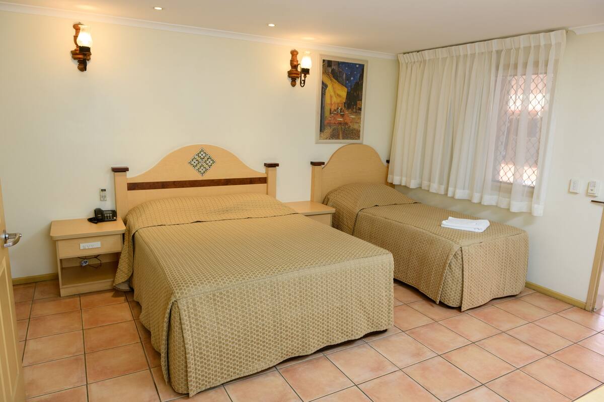 A hotel room with two beds, a nightstand, a lamp, a painting on the wall, and a window with curtains.