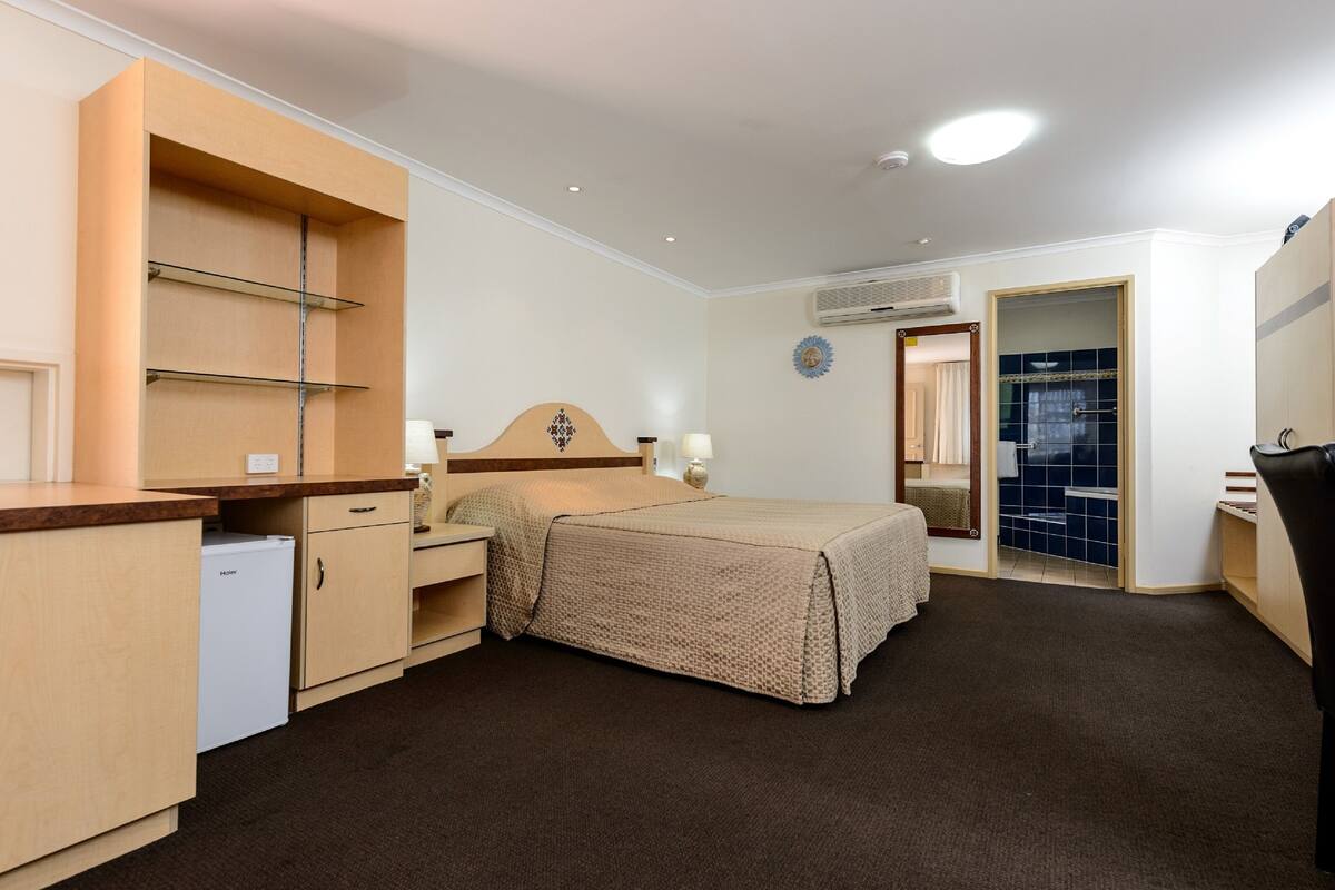 A bedroom with a bed, bedside tables, a wardrobe, a small refrigerator, and an air conditioning unit.