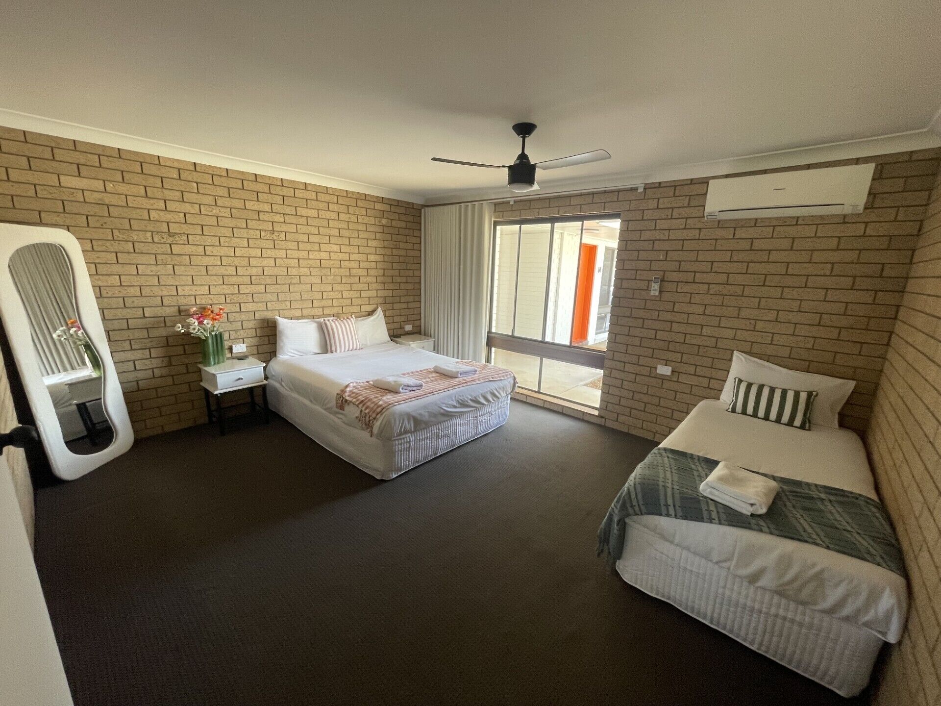 A hotel room with a brick wall, a large bed, a smaller bed, a nightstand with a lamp, a mirror, and a ceiling fan.