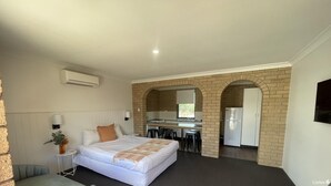 Ein Schlafzimmer mit einem Bett, Nachttisch, Lampe und einer Topfpflanze. Es gibt eine Küchenzeile mit Herd, Spüle und Kühlschrank.
