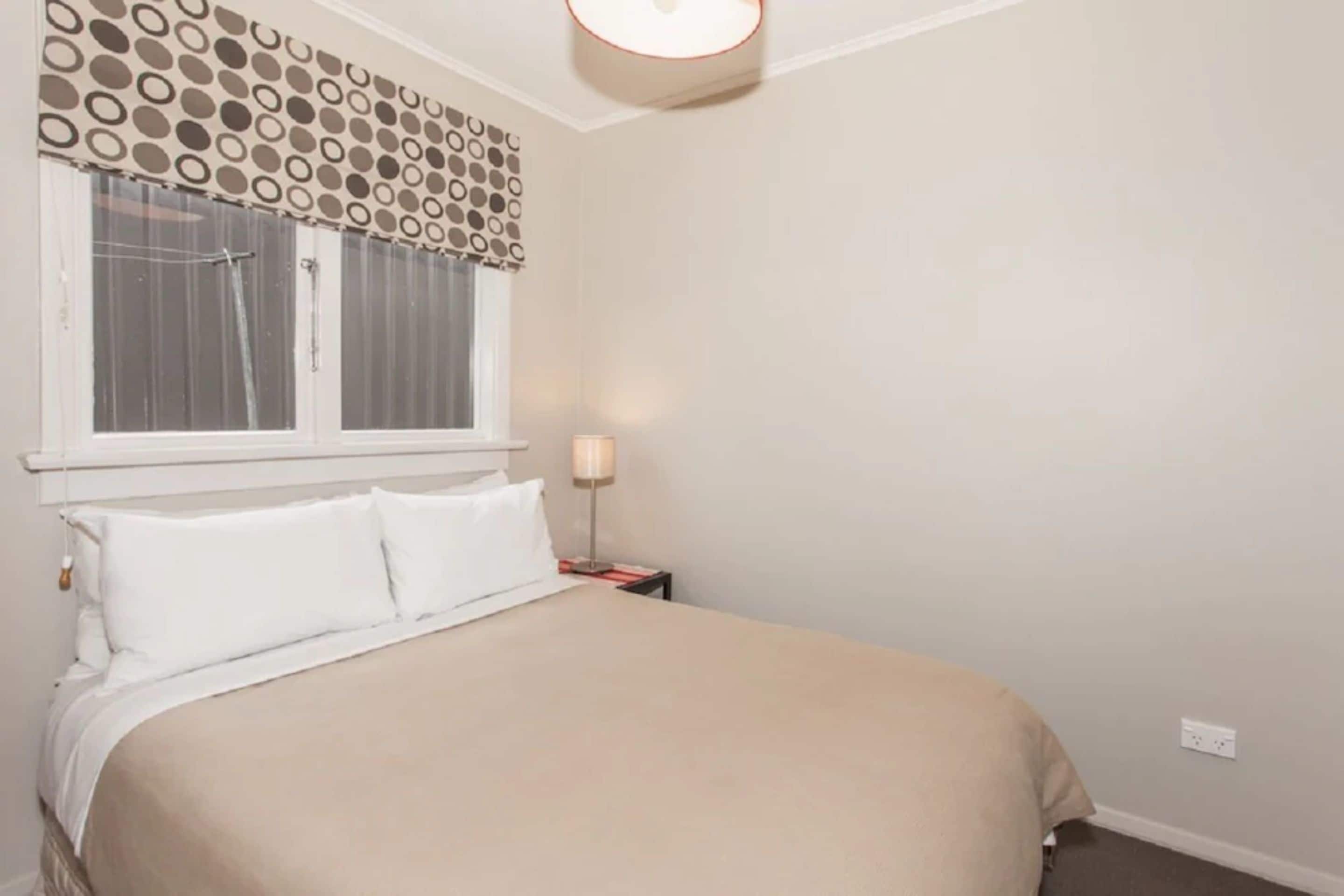 Ein Schlafzimmer mit Bett, weißer Bettwäsche, einem Nachttisch mit Lampe, einem Fenster mit Vorhängen und einer Wand mit einem Rundmuster.