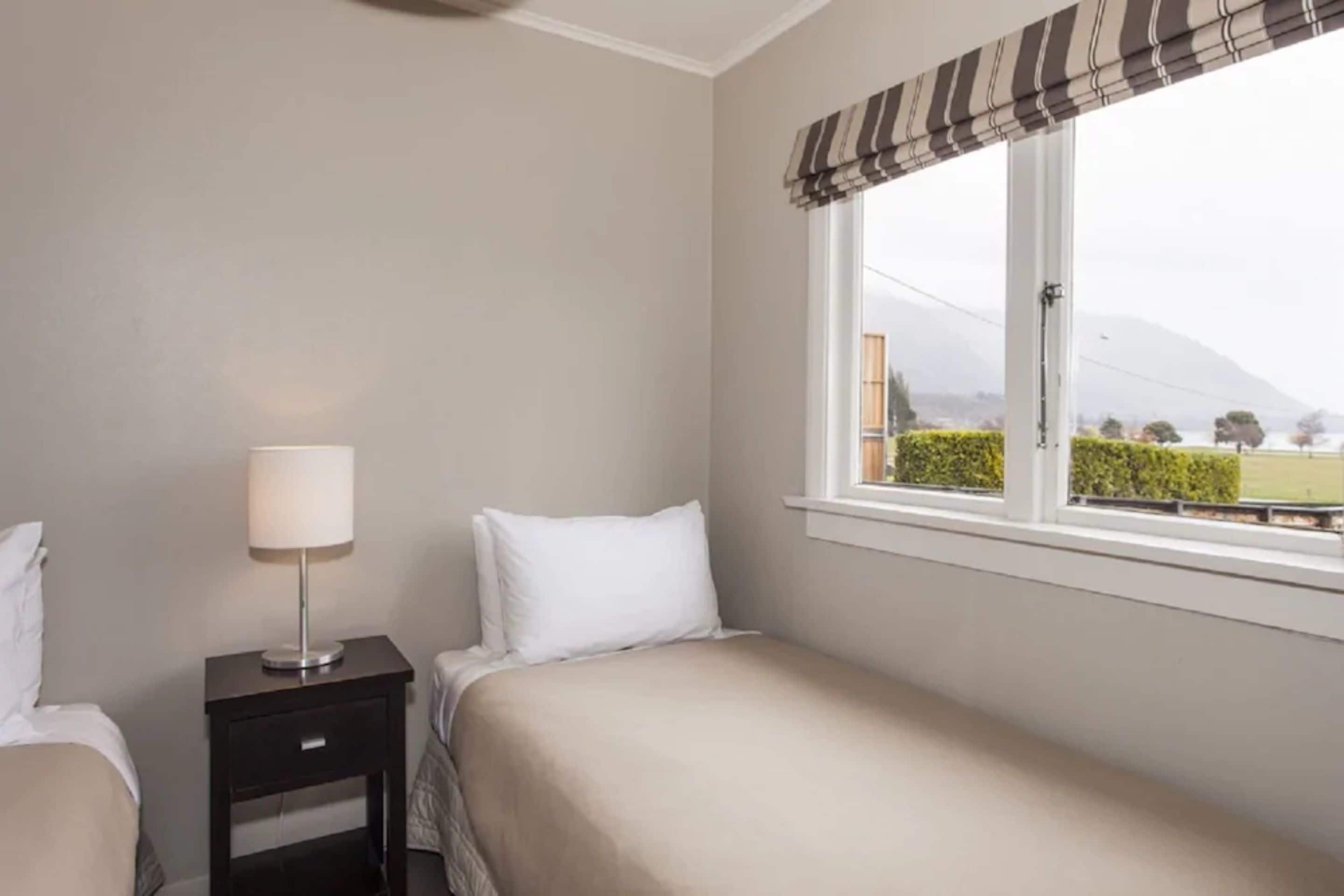 Habitación de hotel con cama, mesita de noche con lámpara y ventana con vista a la montaña y vegetación.