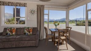 Sala de estar com sofá, mesa de jantar e cadeiras. Há uma janela com vista para montanhas e um céu claro.