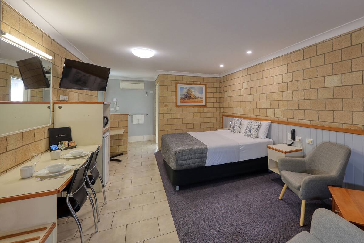 A hotel room with a bed, a desk with a chair, a small table, a telephone, and a picture on the wall.