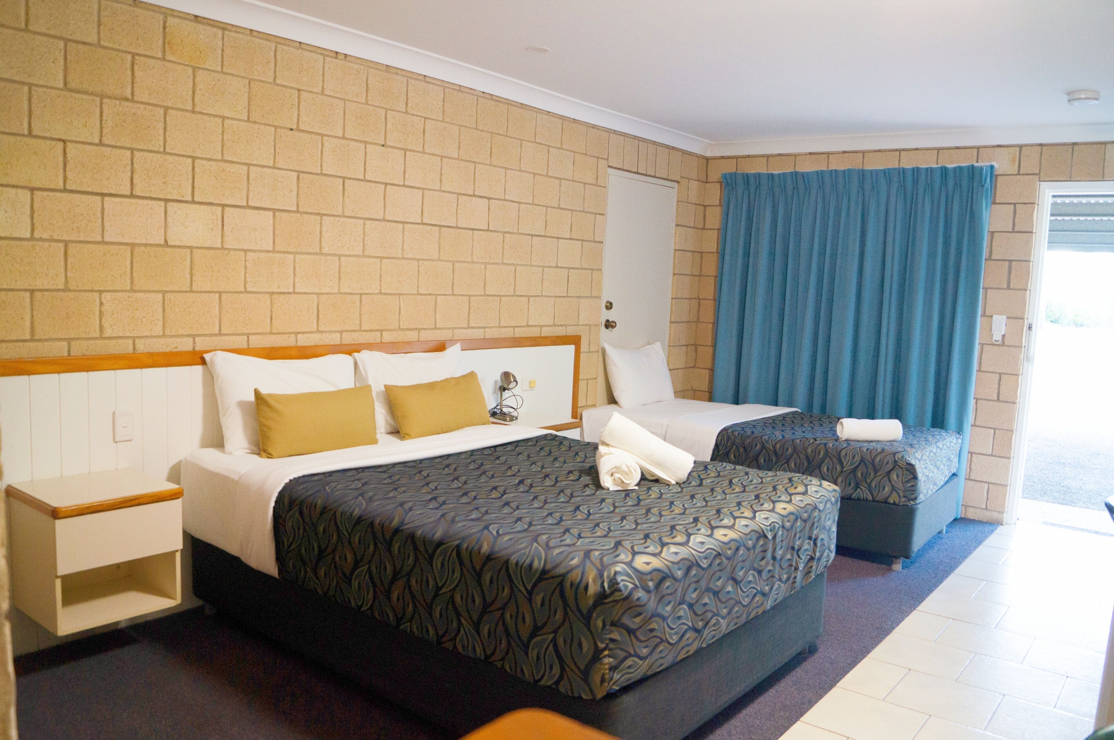 A hotel room with two beds, a brick-patterned wall, a blue curtain, and a white door.