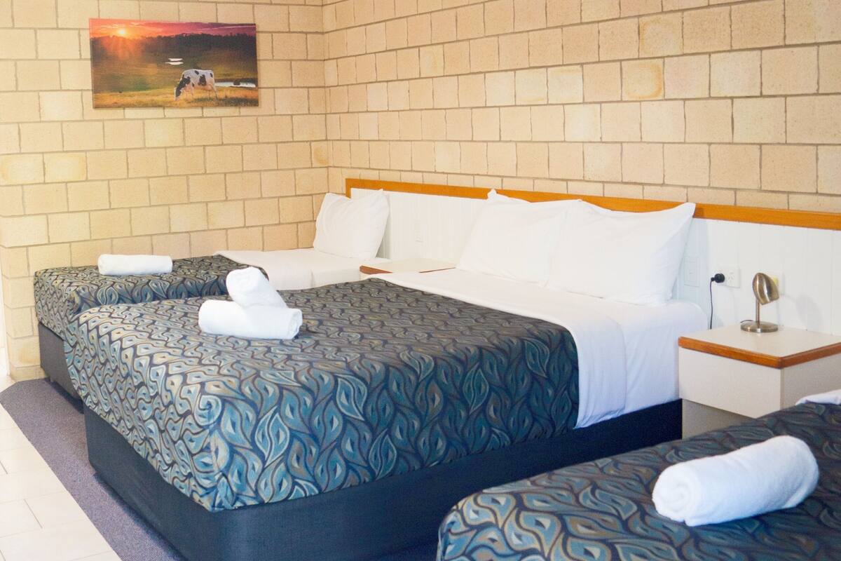 A hotel room with two beds, a brick-patterned wall, a painting of a sunset, and a nightstand with a lamp.