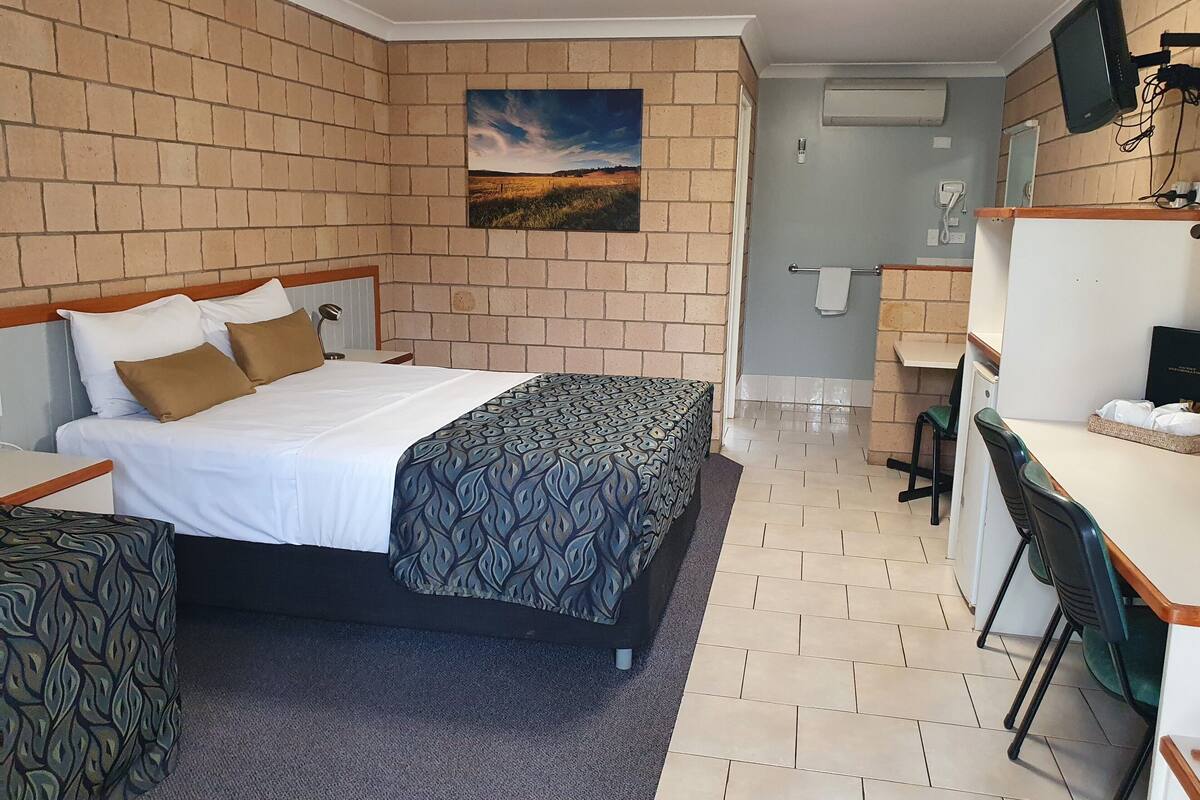 A hotel room with a brick wall, a bed with patterned bedding, a desk, and a small kitchenette.