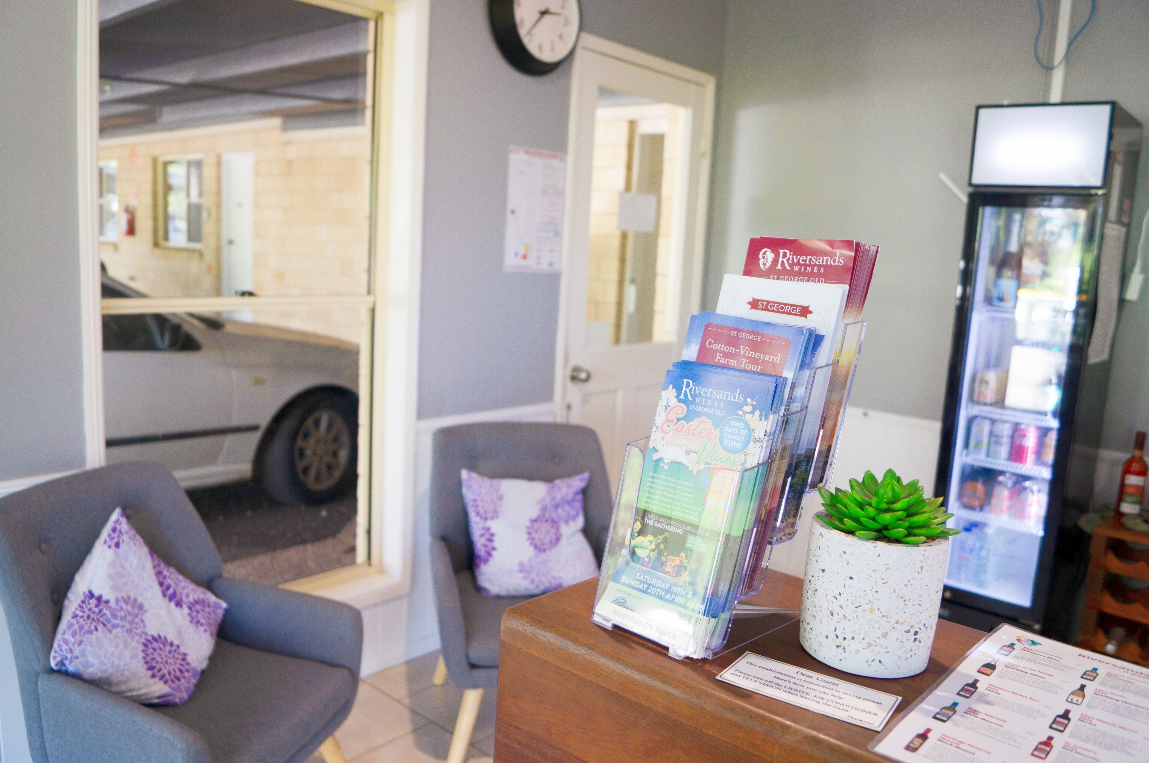 A reception area with a wooden counter, a refrigerator, a clock on the wall, and a car parked outside.