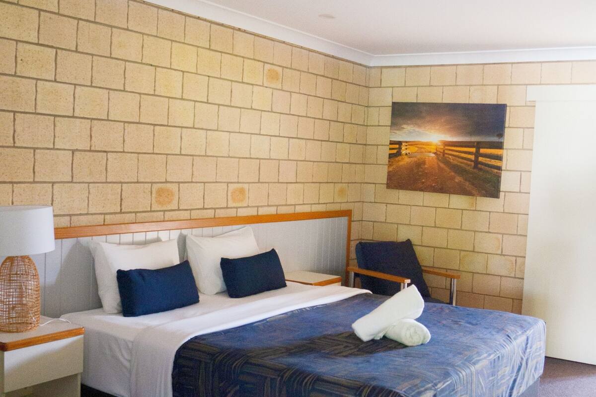 A hotel room with a brick-patterned wall, a bed with a patterned bedspread, a nightstand with a lamp, and a painting of a sunset on the wall.